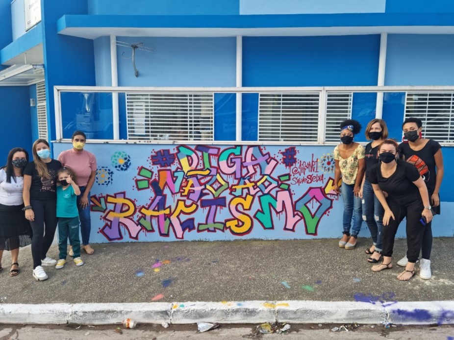 Crianças e adolescentes posam para foto ao lado do grafite feito na frente do Cras Cidade Ademar II, onde se lê a frase “Diga não ao Racismo”