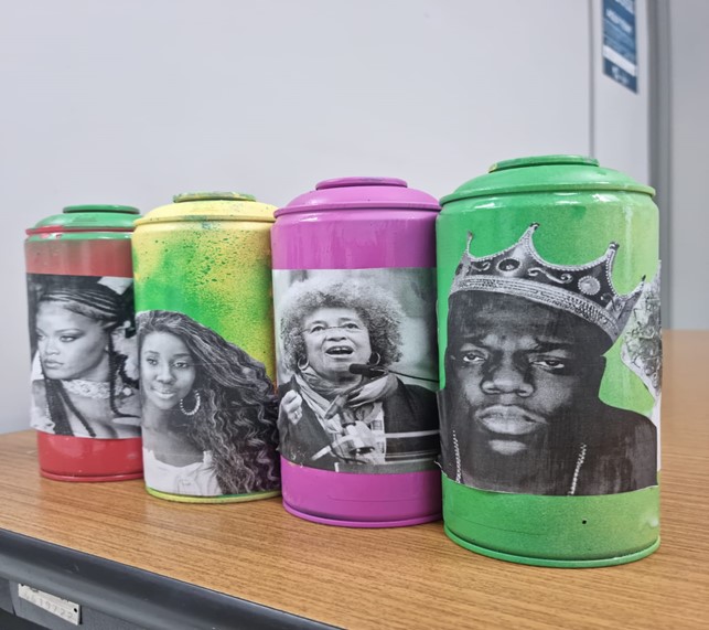 Quatro latas coloridas com grandes figuras em preto e branco: Na primeira lata, da esquerda para a direita, há a imagem da cantora e compositora Rihanna; na segunda lata, vemos Camila de Lucas, influenciadora digital e ex-bbb; na terceira lata está Angela Davis, professora e filosofa integrante do movimento e partido político Panteras Negras. Na quarta lata está a foto do rapper norte-americano The Notorious B.I.G.