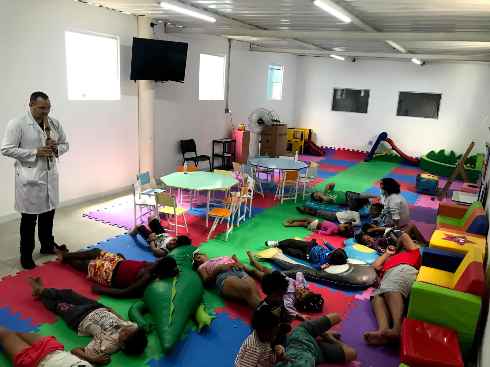 Na brinquedoteca do CTA, diversas crianças estão deitadas nos tatames durante a oficina de musicalidade, um dos passos a serem seguidos. Em pé, o terapeuta toca a flauta doce que rege a atividade. 