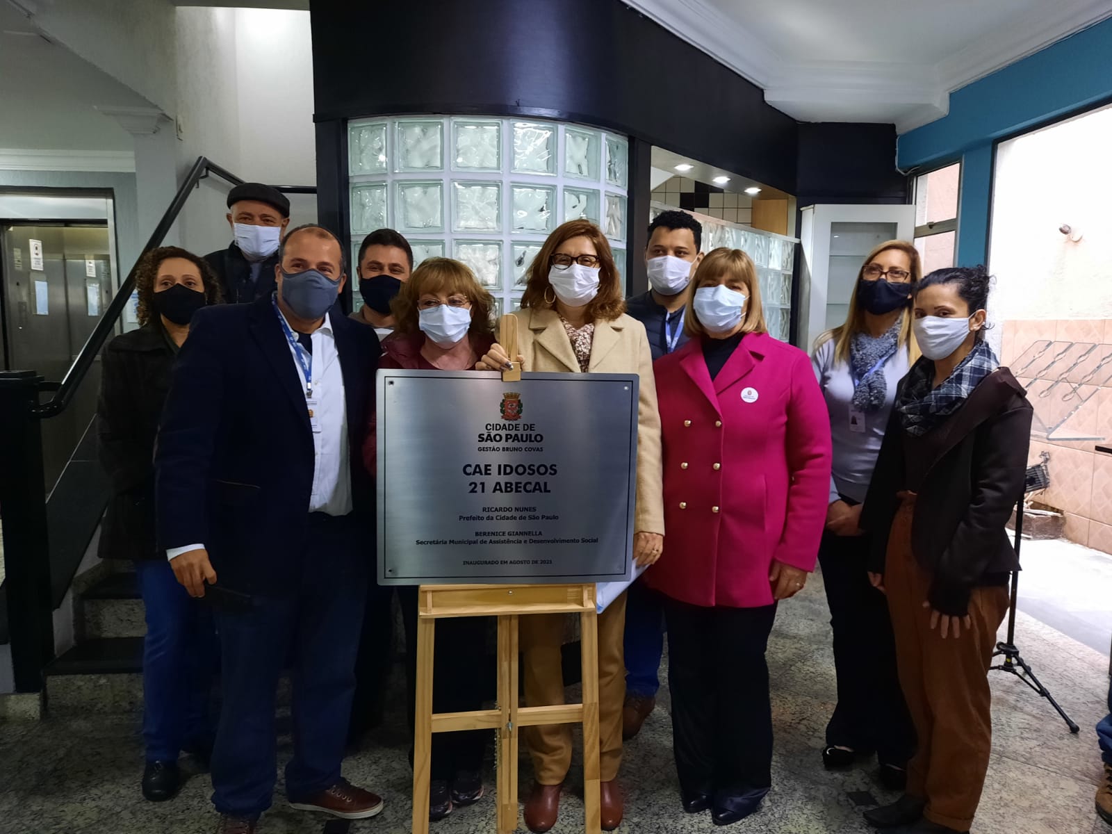 Dez pessoas atrás de uma placa metálica que contém o logotipo da Cidade de São Paulo Assistência e Desenvolvimento Social Gestão Bruno Covas, Centro de Acolhida Especial para Idosos 21 Abecal.