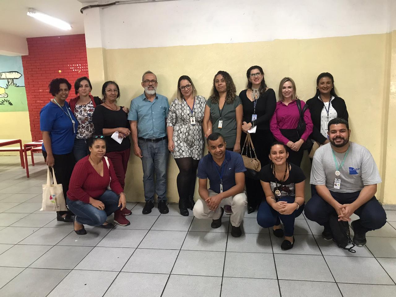Treze pessoas posam para foto em frente a uma parede da cor bege, dividas em duas fileiras, sendo que nove delas estão em pé e outras quatro ajoelhadas.