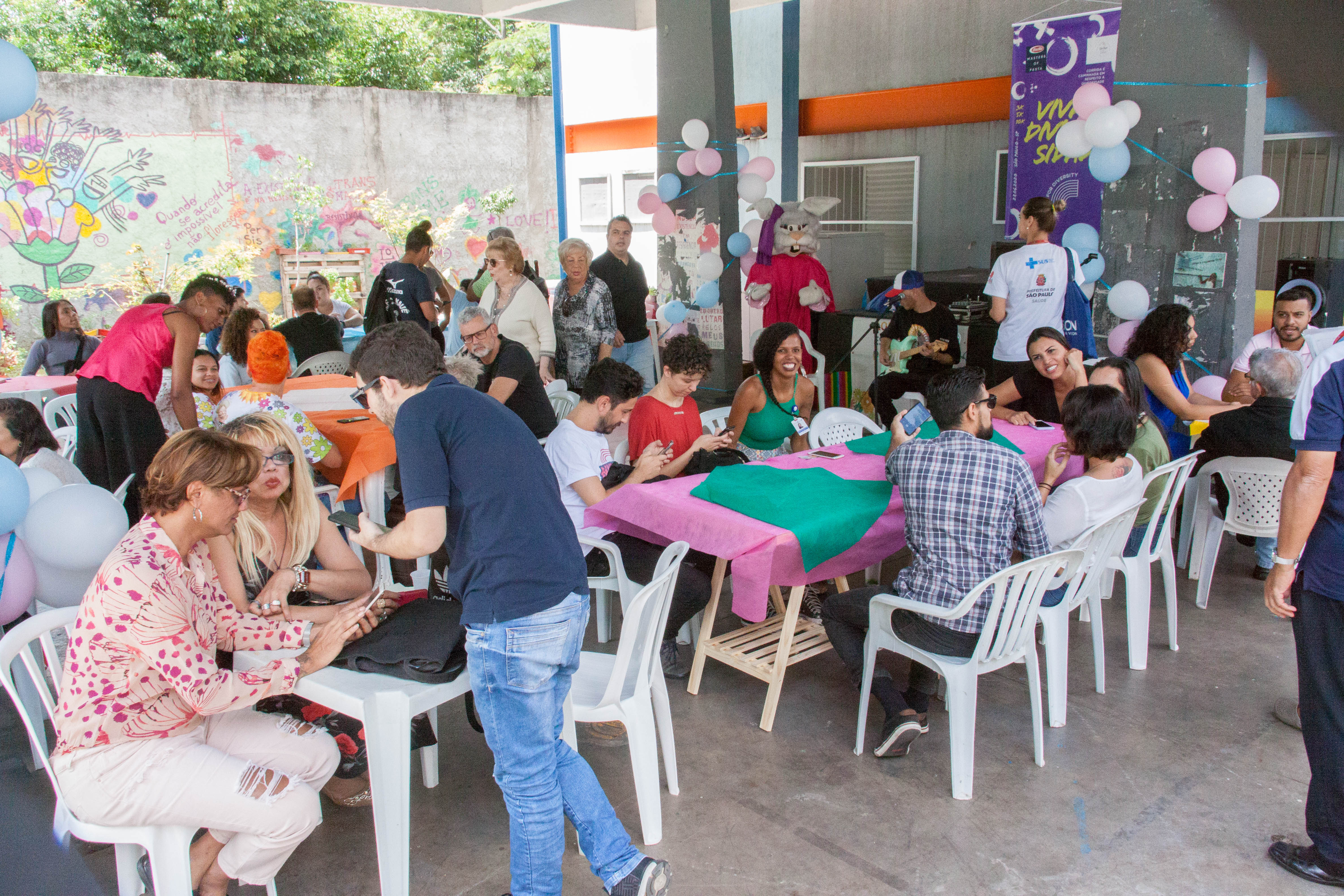 Na parte externa da Casa Florescer, cerca de 100 pessoas estão separadas em diversas mesas decoradas com toalhas amarelas e roxas e sentadas em cadeiras de plástico branco. Todos estão conversando e comendo. O fundo está decorado com balões brancos, rosas e azuis.