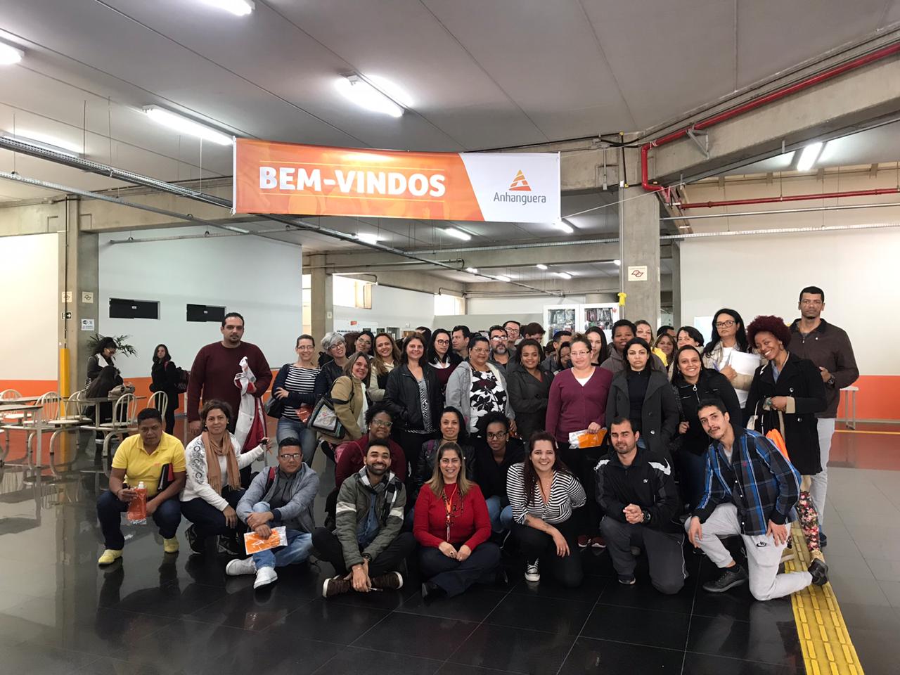 Cerca de 60 pessoas posam para a foto. Acima delas há um cartaz laranja e branco, na parte laranja com letras brancas está escrito bem-vindos. Já na parte branca do cartaz há o logotipo da Faculdade Anhanguera.