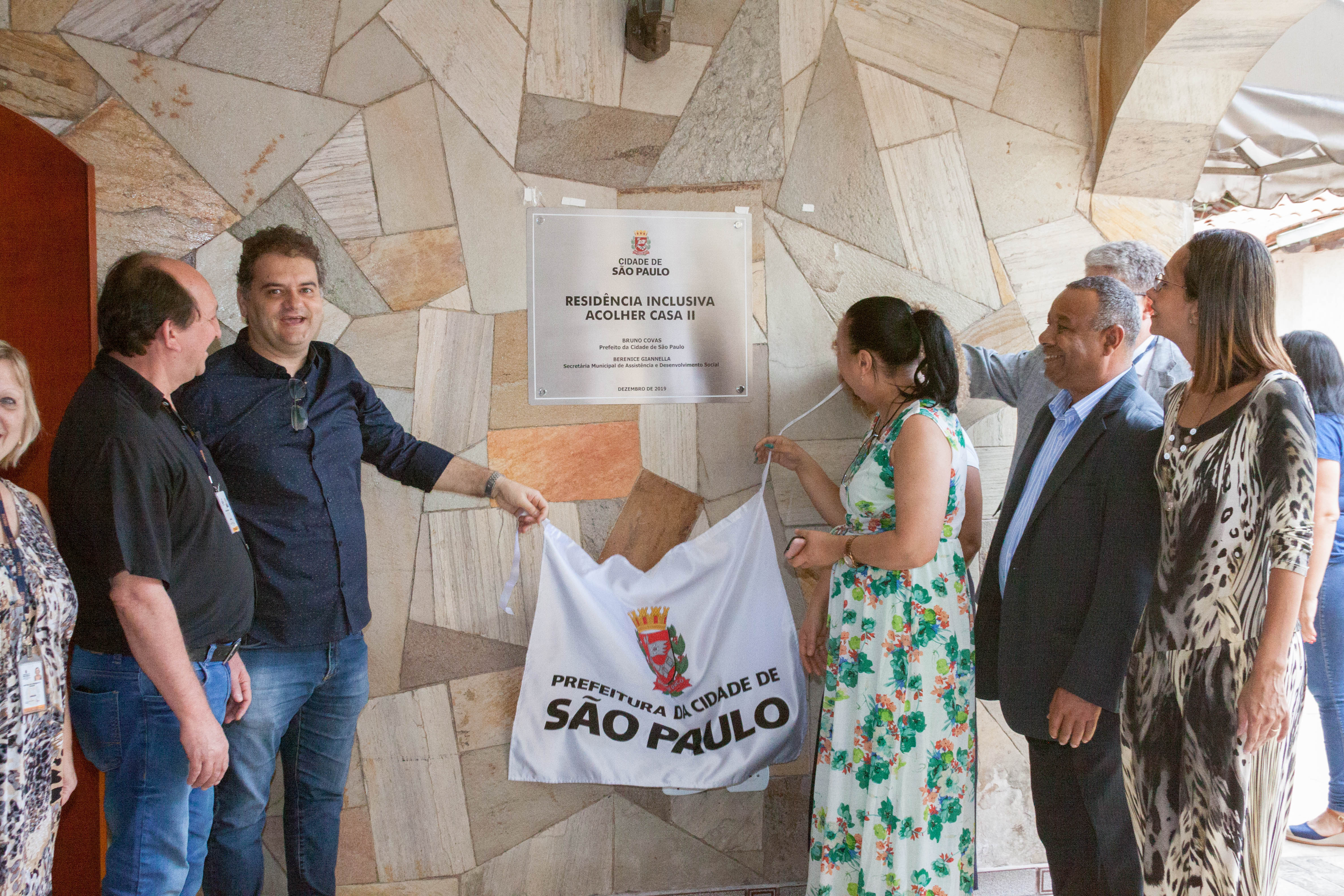 Em parede de pedras marrons, uma placa cinza está grudada na parede. Na placa há o logo da Cidade de São Paulo e abaixo em letras pretas está escrito “Residência Inclusiva Acolher Casa Dois”. A frente da placa, há sete pessoas em pé e duas delas removendo um pano branco que também contém o logo da cidade.