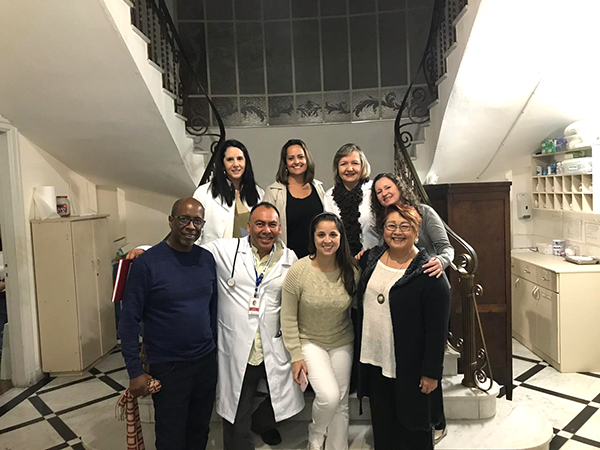 #PraCegoVer: Equipe da hospedaria composta por oito pessoas, divididas em duas fileiras, posam para a foto em frente a uma escada do local