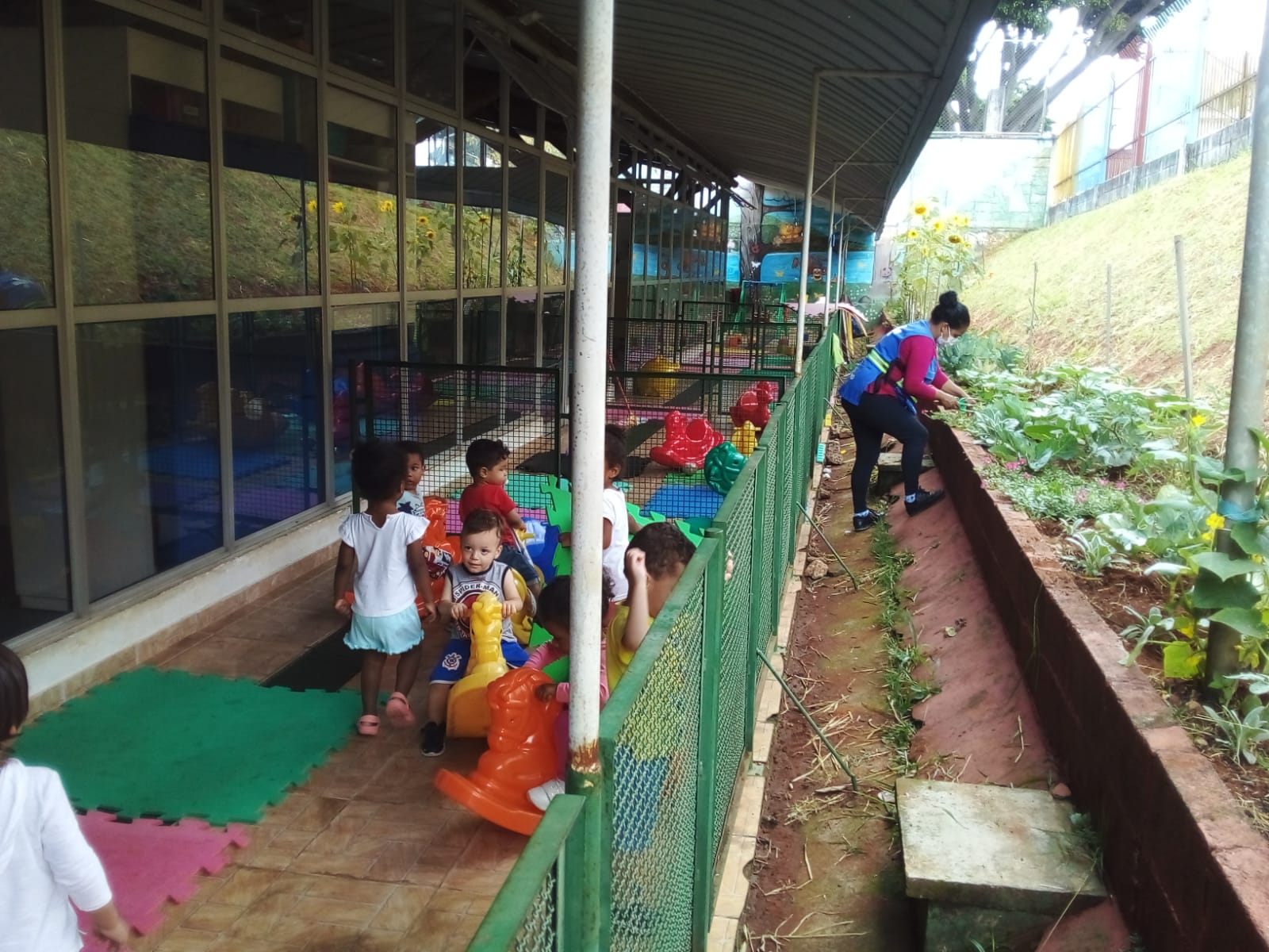 A foto mostra, do lado esquerdo, um grupo de crianças pequenas brincando em um pátio coberto e cercado; à direita, do lado de fora, uma mulher vestindo um colete azul cuida de uma horta com várias espécies vegetais, plantada em um canteiro lateral