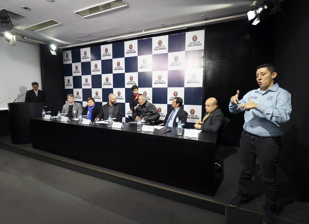 mais de dez pessoas no palco, na sala de imprensa da Prefeitura de São Paulo. Ao lado do Prefeito Bruno Covas, o secretário municipal da Pessoa com Deficiência, Cid Torquato, fala ao microfone. 