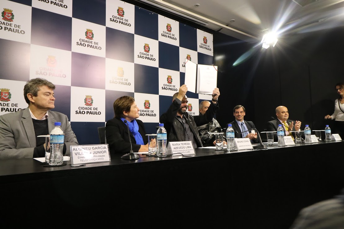 atrás da mesa, no palco, mais de dez pessoas. Prefeito Bruno Covas levanta o termo do contrato assinado, sorrindo. 