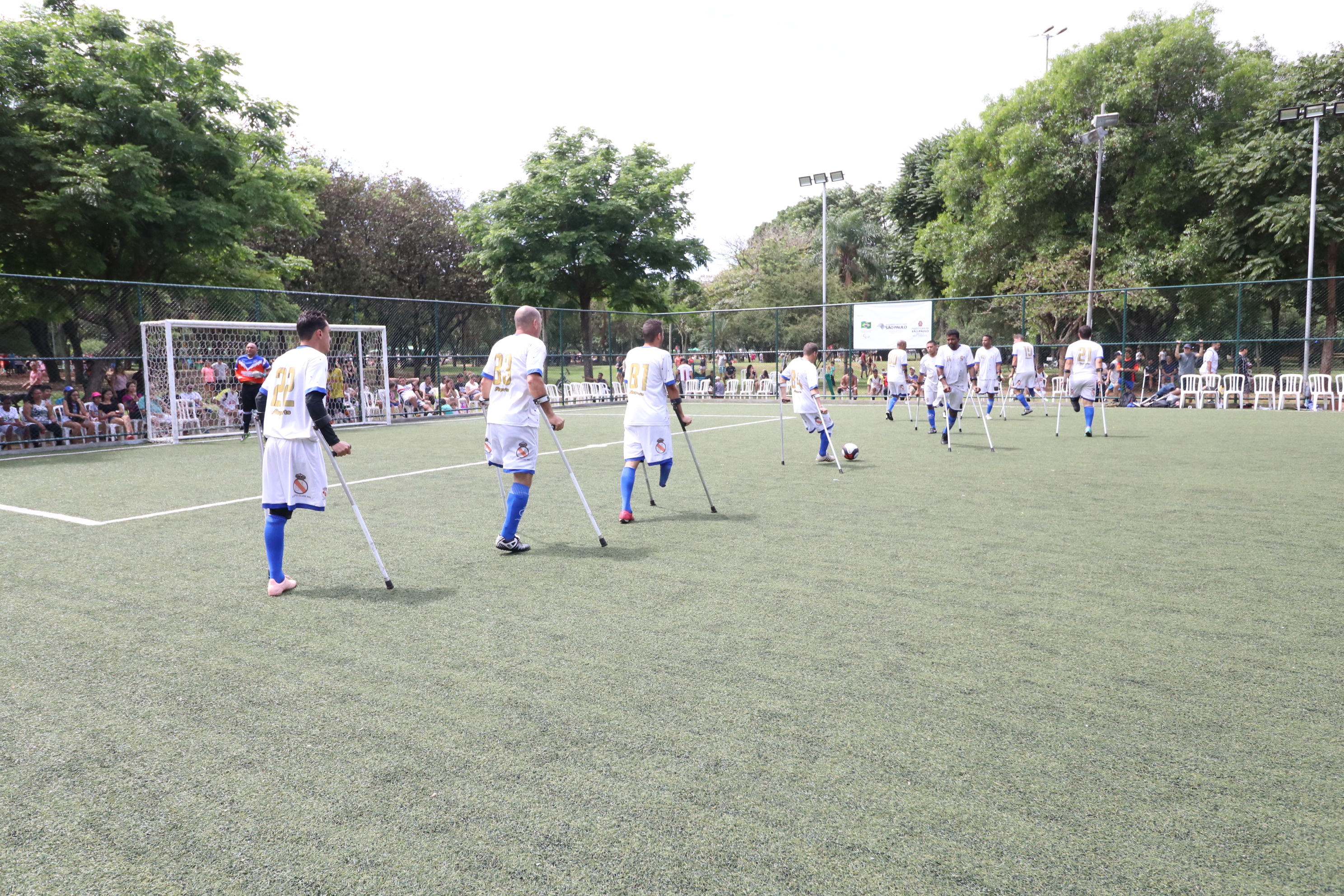 Jogadores amputados durante competição. Campo de futebol é aberto com gramado e árvores ao redor. 