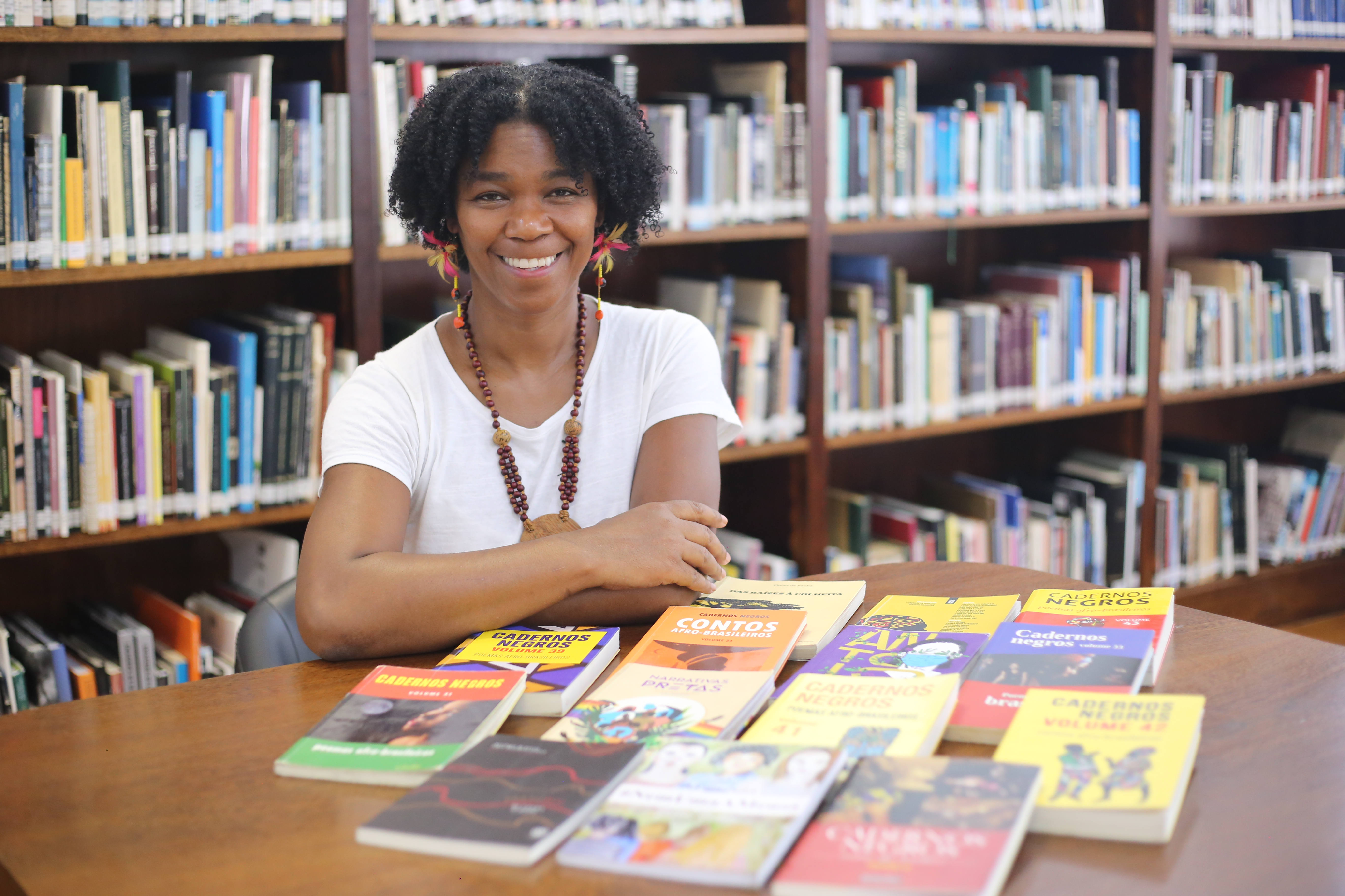 A imagem mostra a personagem, Claudia Walleska, uma mulher negra com cabelos crespos e curtos, vestindo uma camiseta branca, usando um colar e  brincos coloridos. Ela está sentada em uma mesa com livros e uma estante ao fundo, também com diversos livros