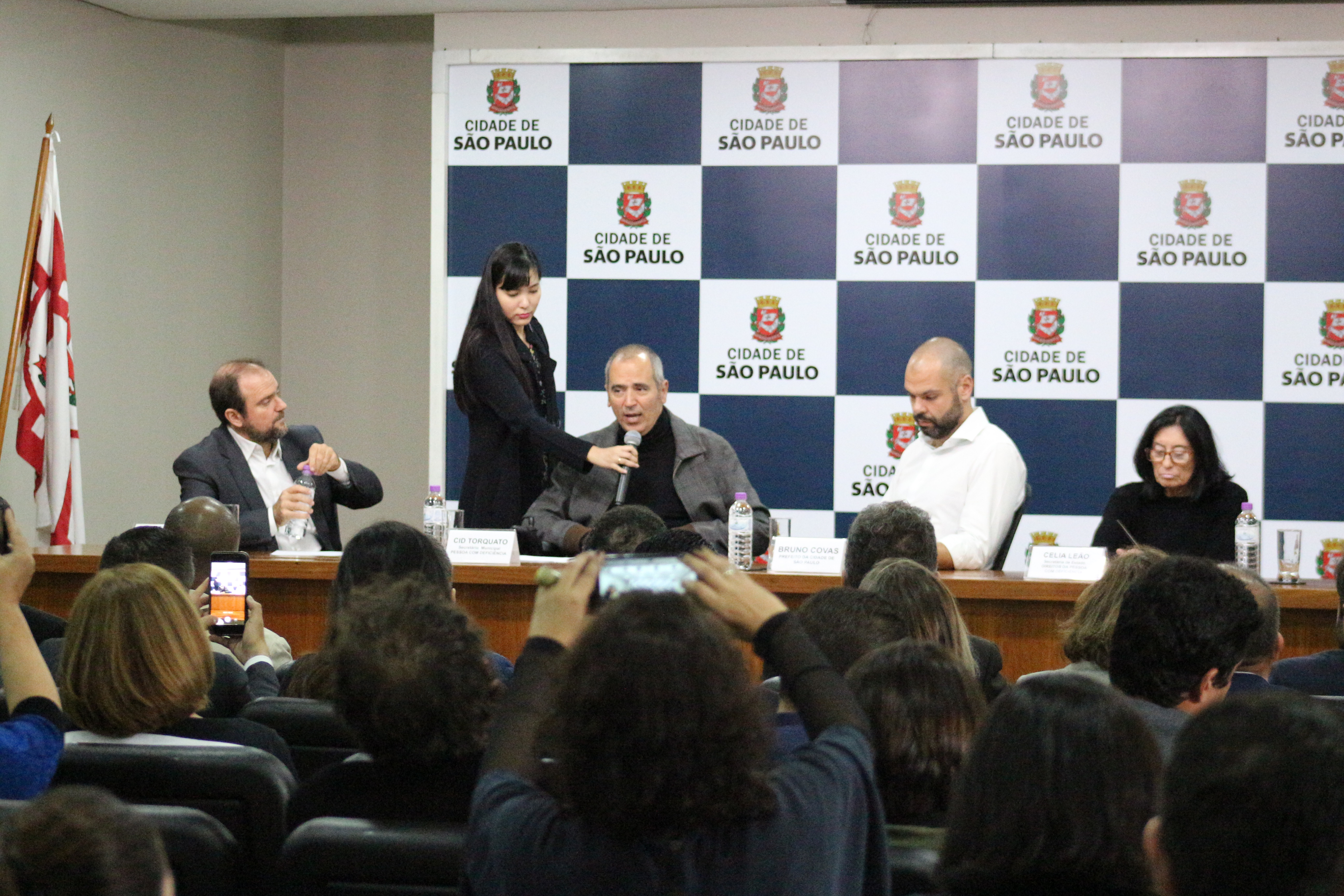 foto panorâmica da mesa de abertura com prefeito Bruno Covas e secretários. Mais de trinta pessoas na foto. Secretário Cid Torquato fala ao microfone. 