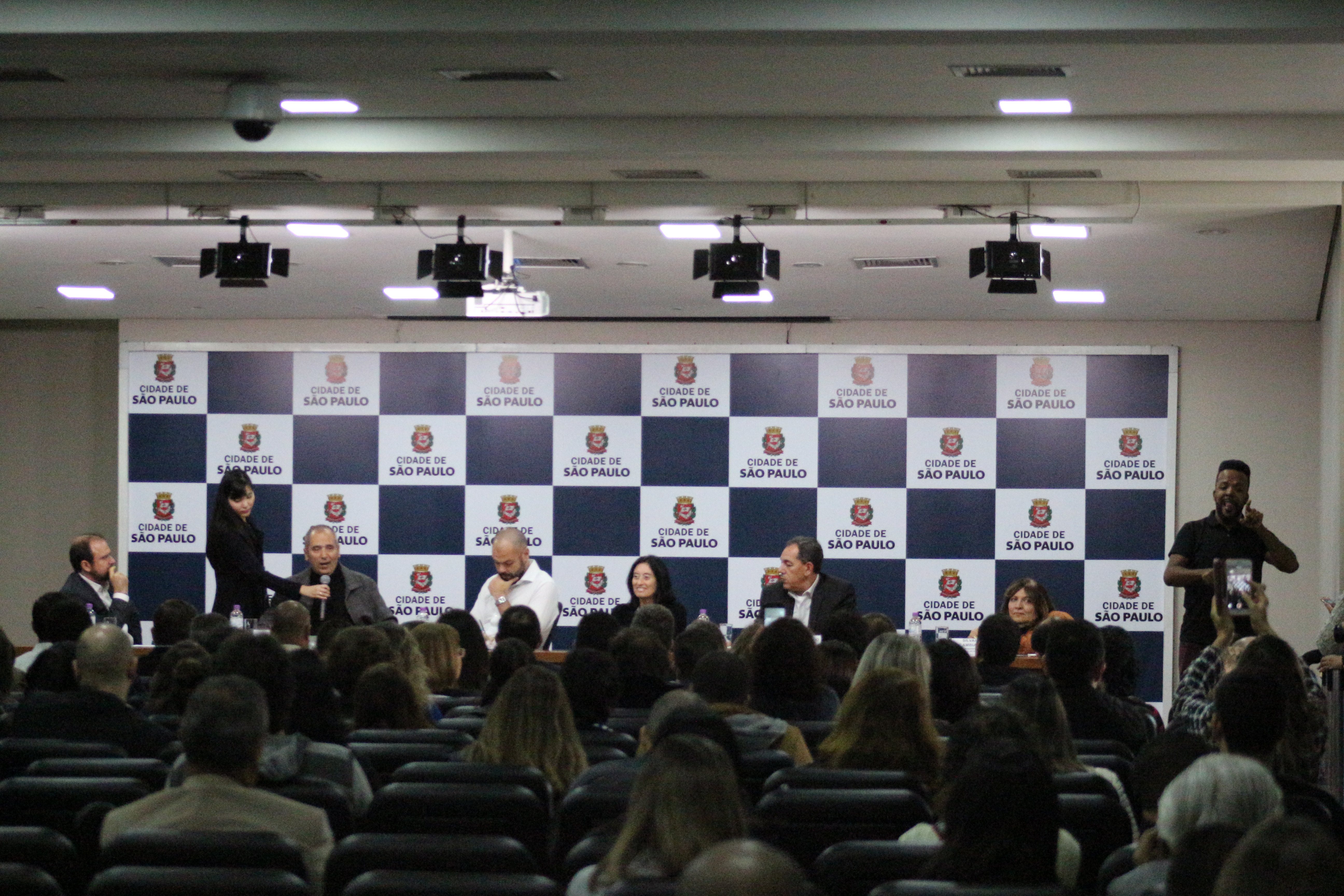 foto panorâmica da mesa de abertura com prefeito Bruno Covas e secretários. Mais de trinta pessoas na foto. Secretário Cid Torquato fala ao microfone. 