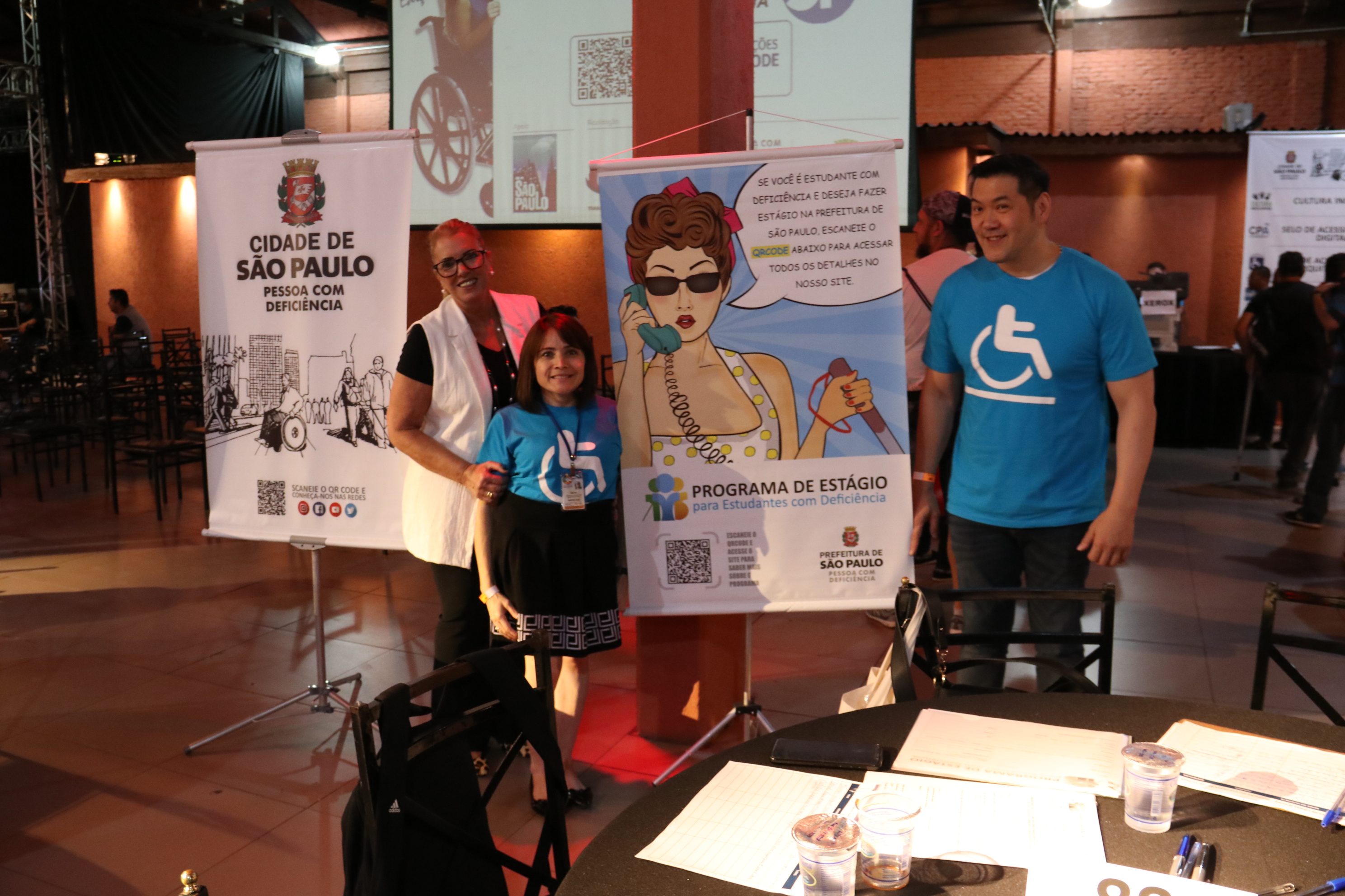 três pessoas na foto, próxima do cartaz  e mesa da SMPED, a secretária adjunta Marinalva Cruz. 