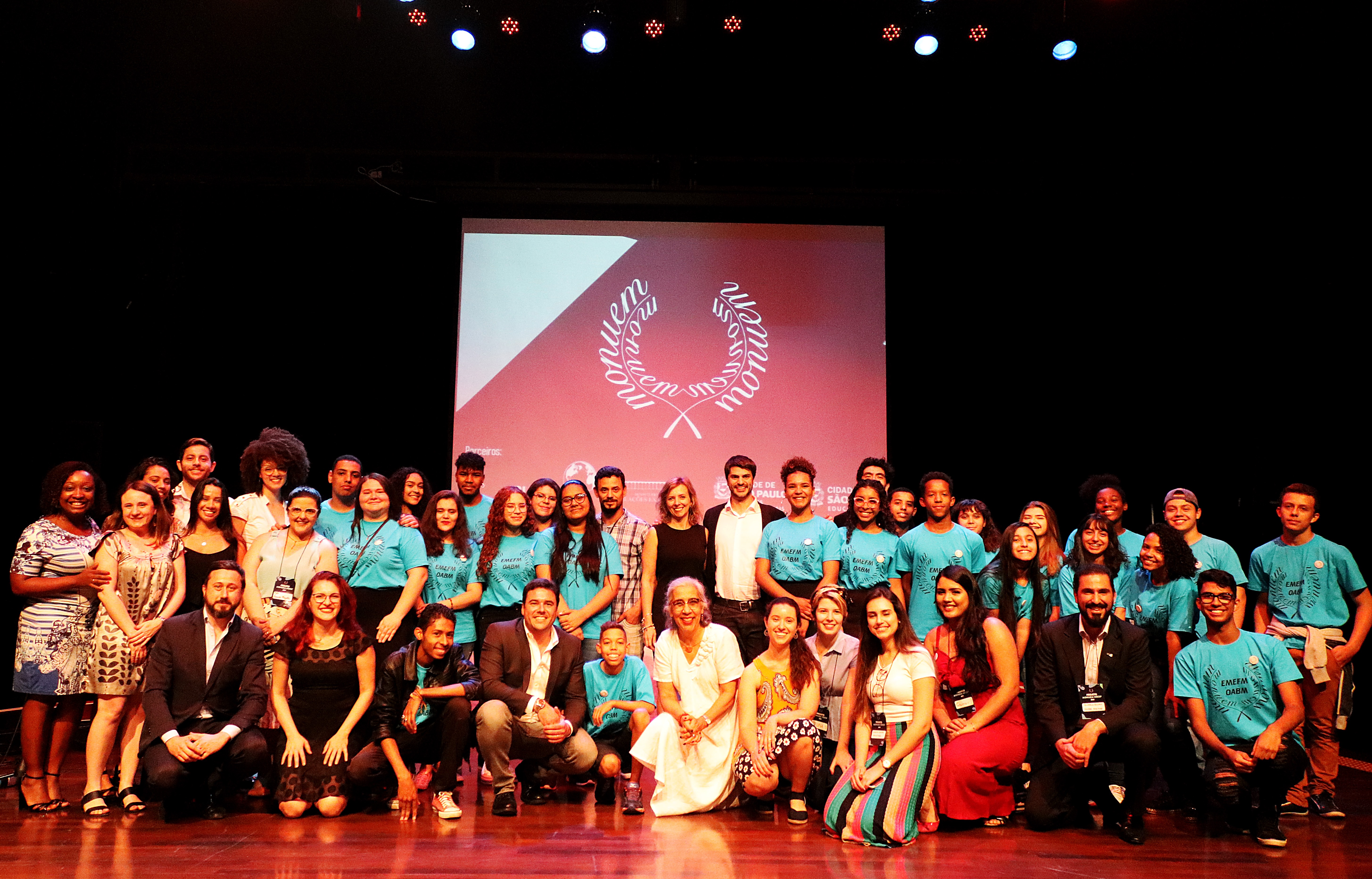 Participantes do Projeto posam para foto em cima do palco. O fundo é preto, há um telão com o logotipo do Monuem em branco e vermelho, os estudantes vestem camiseta azul.