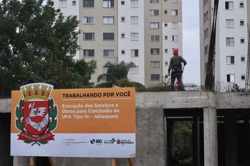 #PraCegoVer: Uma placa na cor laranja sinalizando que ali é uma obra da UPA Jabaquara. De fundo temos um prédio nas cores bege e branco. Podemos observar um trabalhador da obra, com capacete vermelho.um 