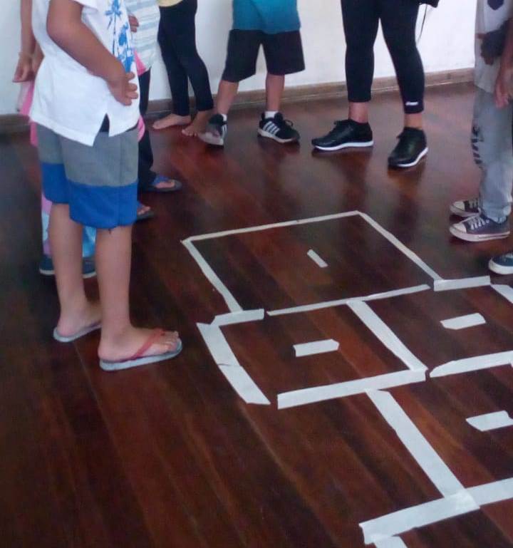 Chão marrom de uma quadra onde estão marcações feitas com fita branca para poder brincar de ‘amarelinha’. Na mesma foto, crianças aparecem da cintura pra baixo, ao redor da marcação. 
