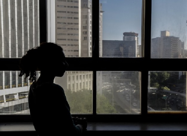 #PraCegoVer: Foto1: Em uma grande janela, há uma uma mulher de costas olhando a paisagem de uma avenida de São Paulo, composta por prédios e arvores ao redor.