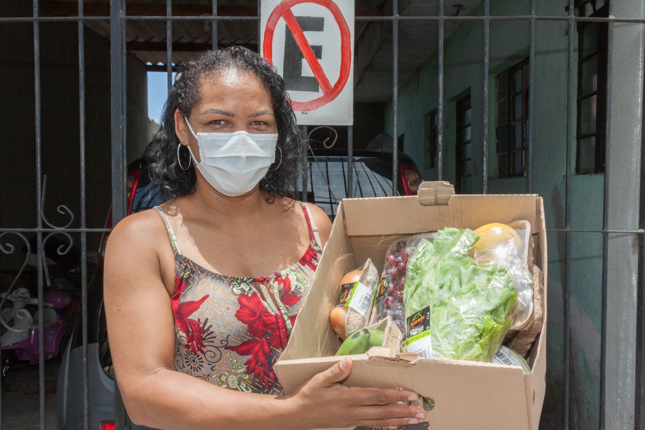 Uma mulher com máscara de proteção facial posa para foto e segura uma cesta de alimentos agroecológicos e está em frente a sua residência. 