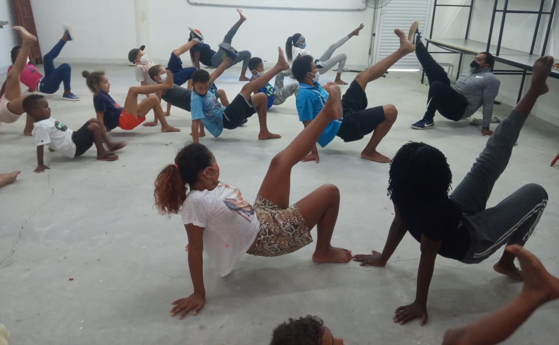 Em uma sala com o piso claro, diversas crianças estão apoiadas sobre suas mãos, com apenas um pé no chão e com o tronco voltado para cima. O exercício feito é guiado pelo professor que está no canto superior direito da foto. 