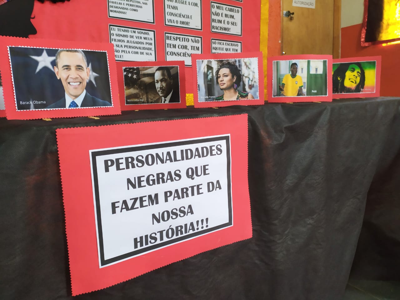 Mesa que expõe retratos de personalidades negras, na parede de fundo aparece diversas frases de desconstrução e engajamento da causa racial.