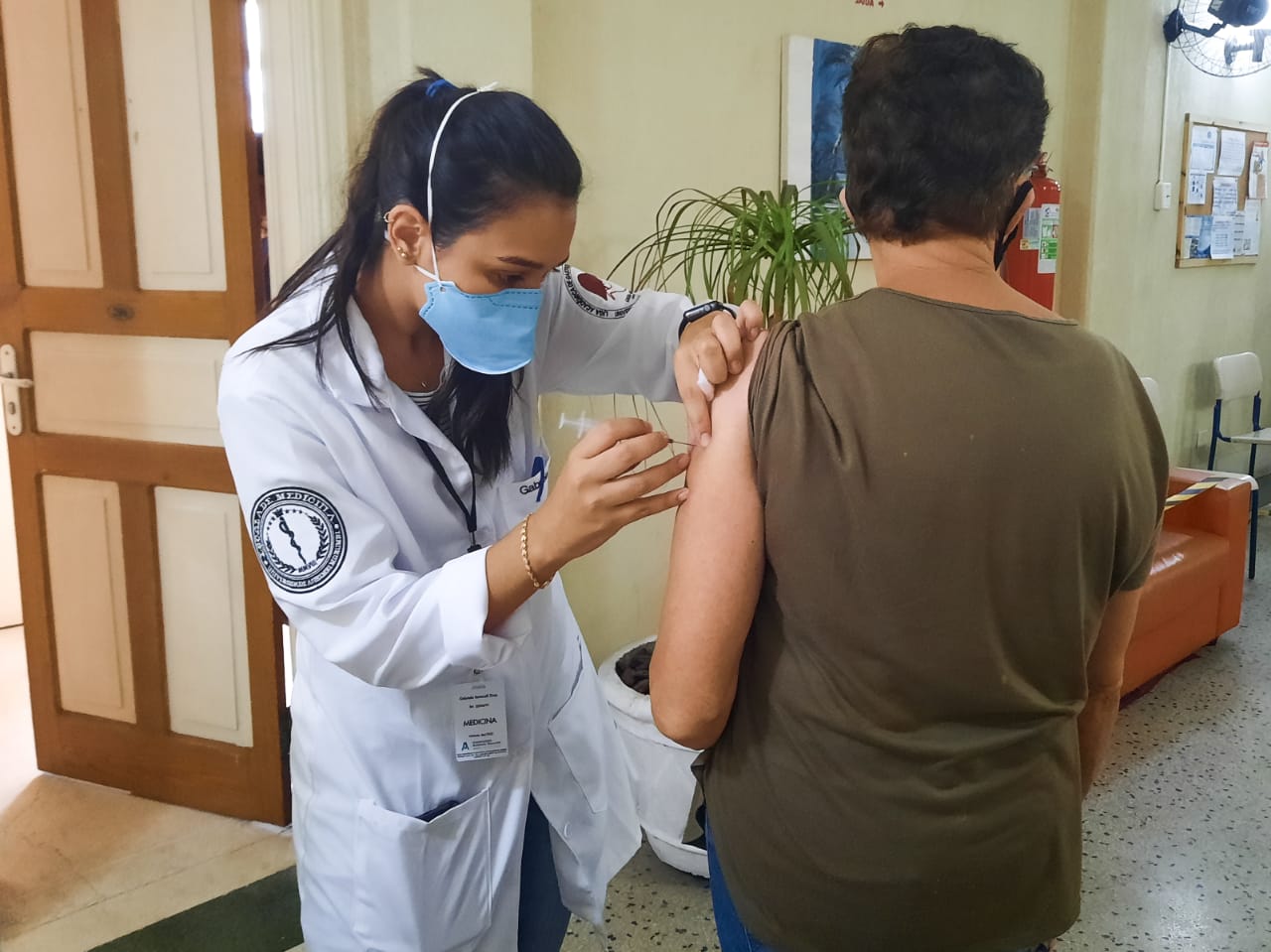 Enfermeira aplica vacina em uma senhora de cabelos curtos que está posicionada de costas para a câmera, recebendo a vacina no braço esquerdo.