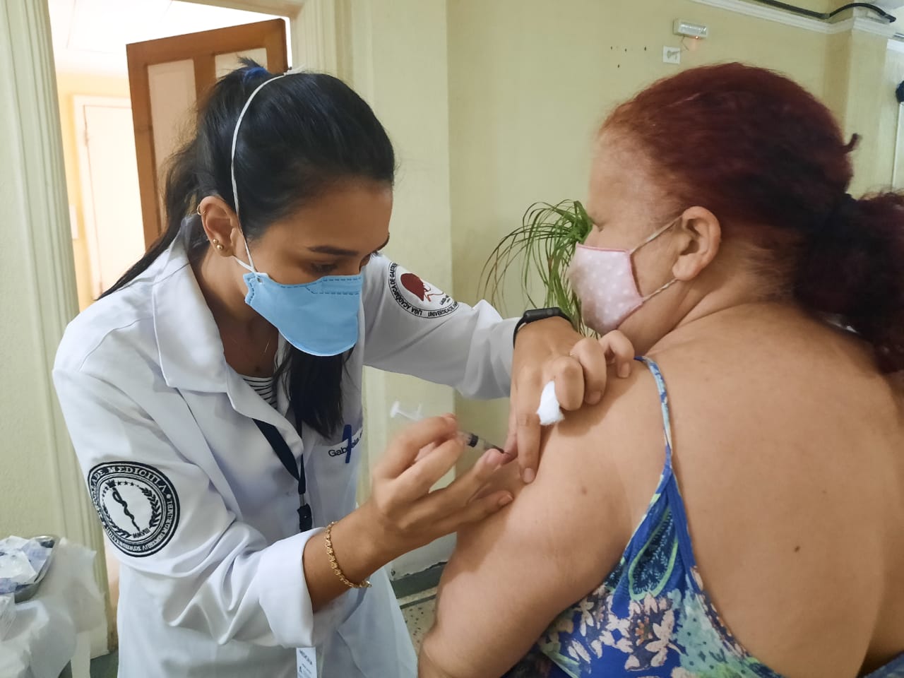 Enfermeira aplica vacina em uma senhora de cabelos ruivos que está posicionada de costas para a câmera, recebendo a vacina no braço esquerdo. 