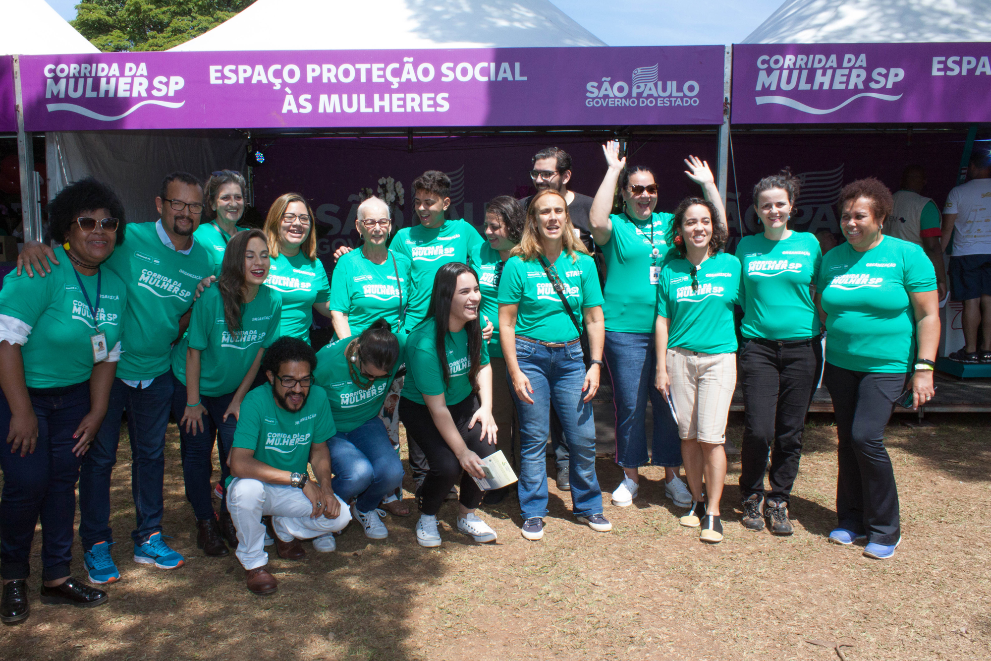 17 pessoas vestidas com blusas verdes sorriem para a foto em frente ao Espaço Proteção Social às Mulheres.
