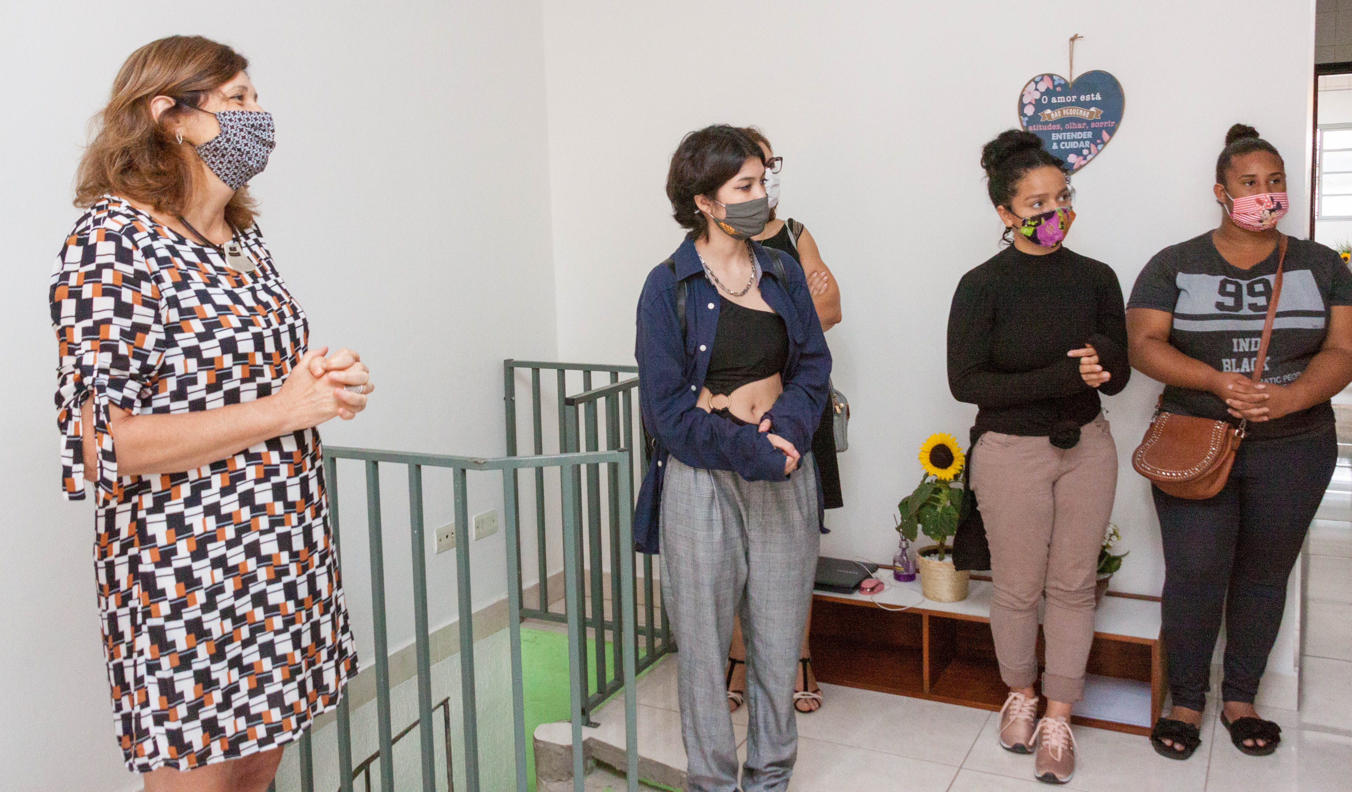 A secretária Berenice e as três moradoras da casa estão em pé na sala de estar da República, um ambiente todo branco com um coração na parede e um vaso de girassol em cima de um aparador baixo. À esquerda, grades de proteção de cor verde limitam o acesso ao piso inferior da casa.