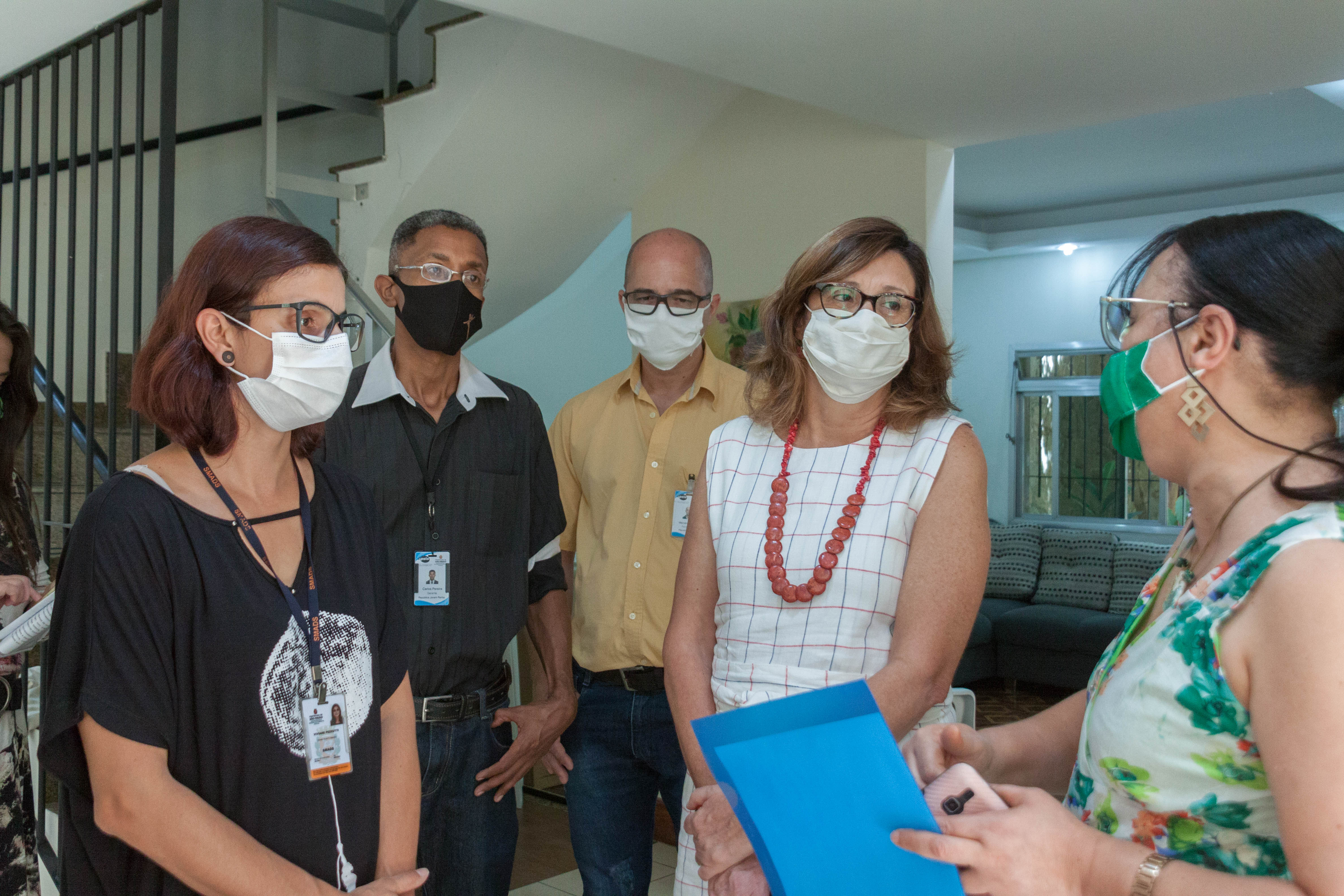 A supervisora de Assistência Social da região da Penha e a equipe técnica da casa recebem a secretária Berenice Giannella. São cinco pessoas na imagem, três mulheres e dois homens que conversam em pé, em frente a escada de acesso ao piso superior da casa.