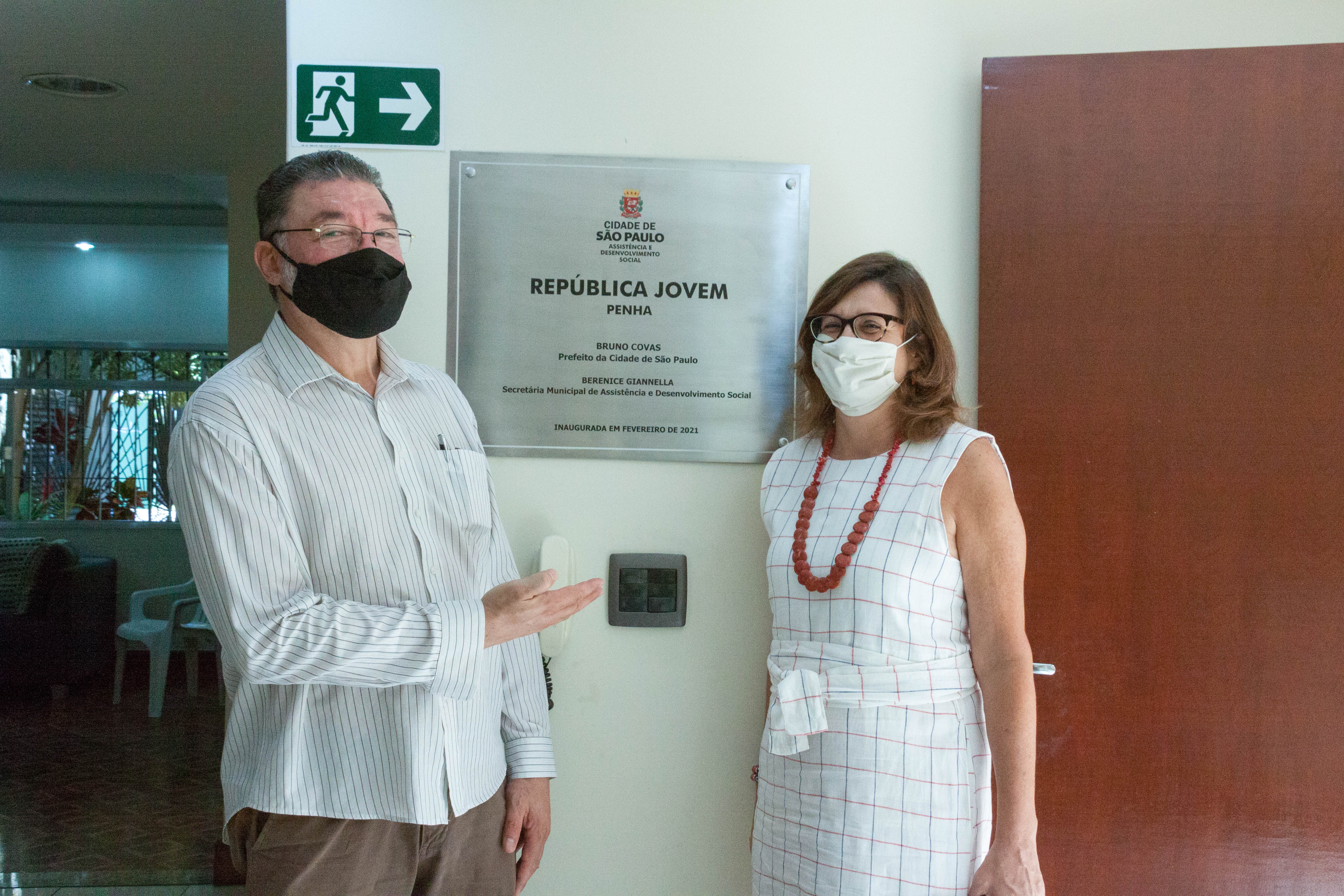 O presidente da Associação Apoio e a secretária Berenice Giannella estão posicionados à frente da placa de inauguração da casa. A placa é de aço escovado, com brasão da Prefeitura e o nome do equipamento: República Jovem Penha, escrito em letras pretas.