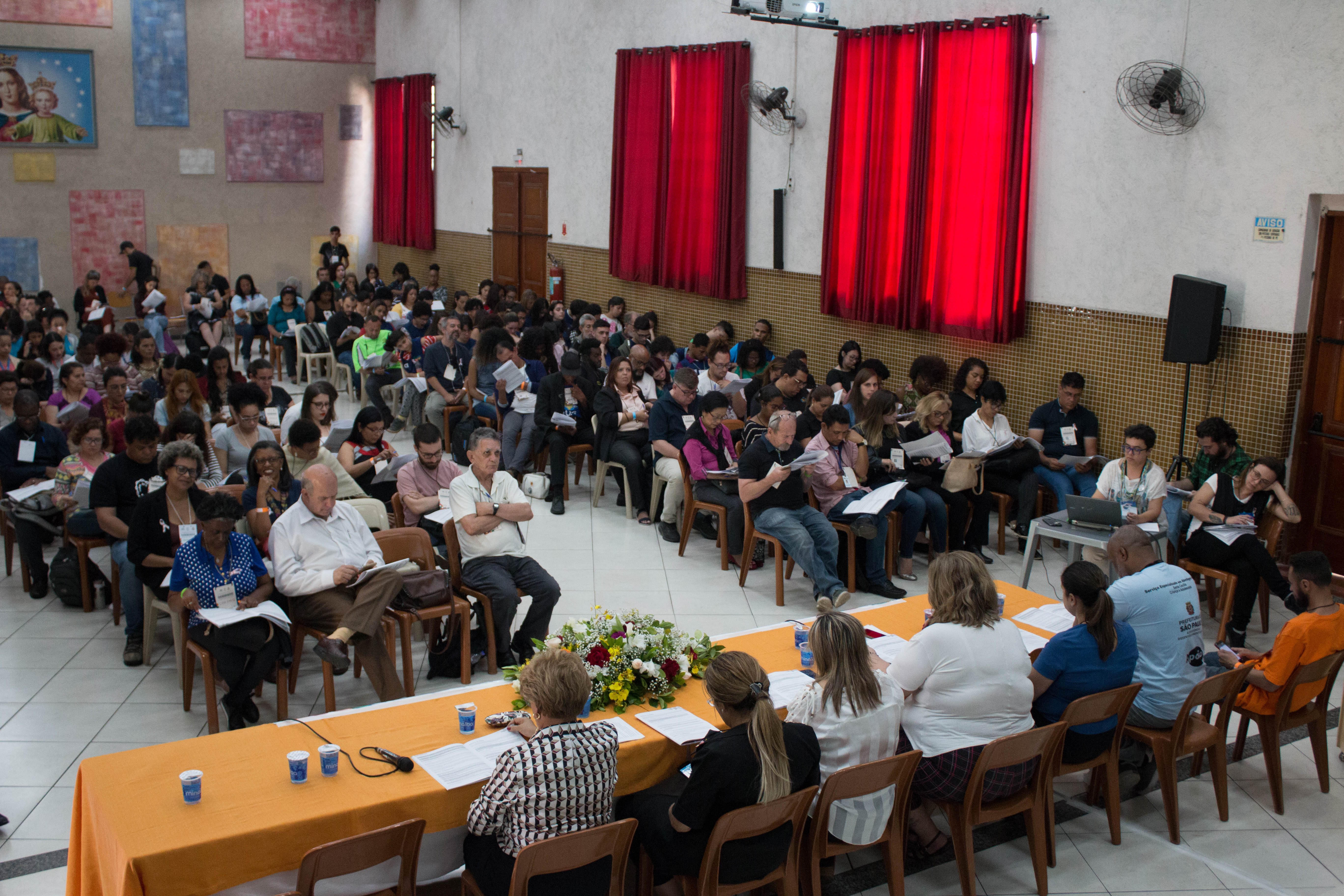 Um salão com várias pessoas discutindo temas que envolvem políticas públicas para assistência social.