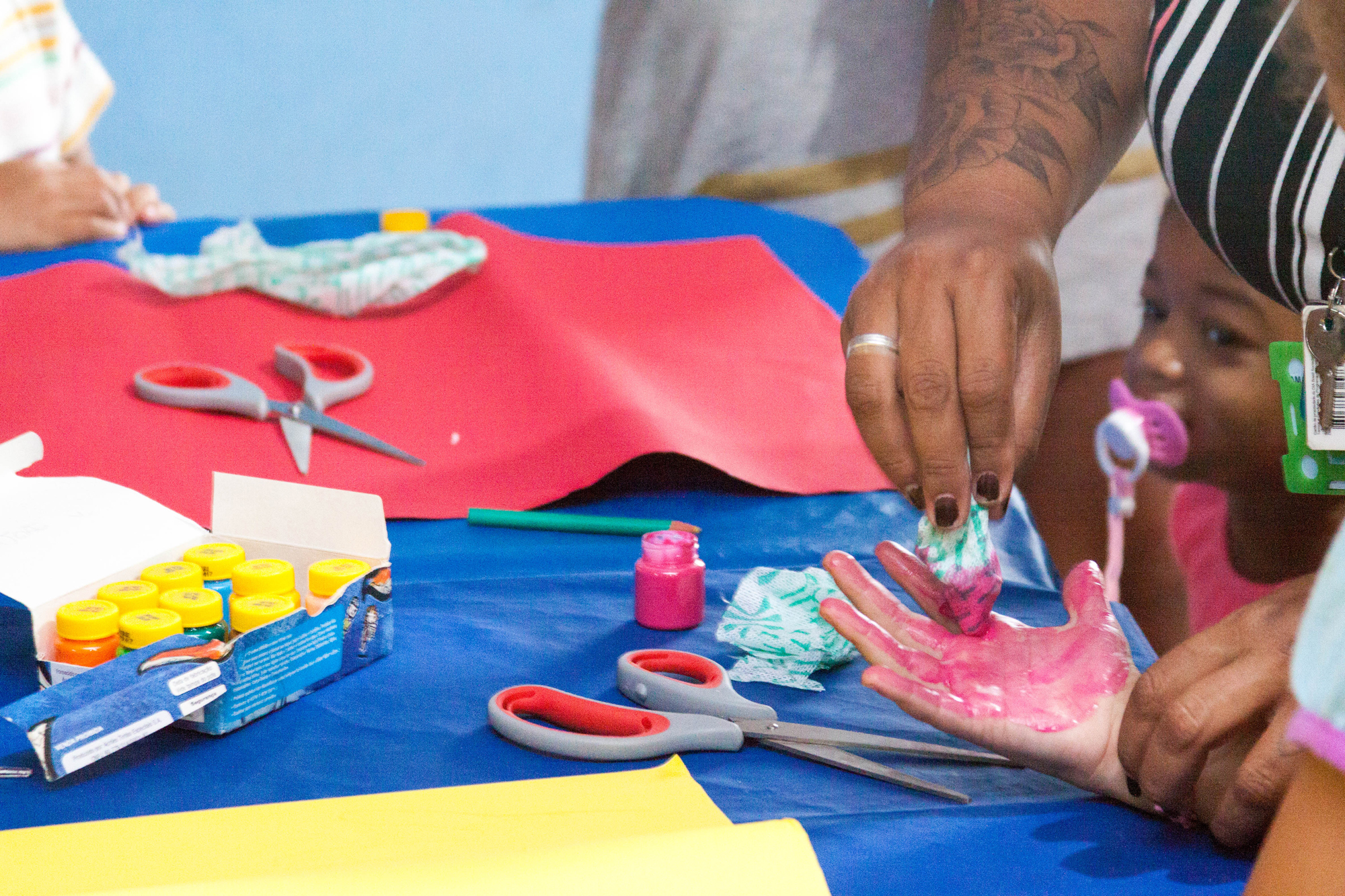 Uma mesa repleta de cartolinas coloridas e tintas guache. Há uma mão feminina auxiliando a pintura da mão de uma criança.