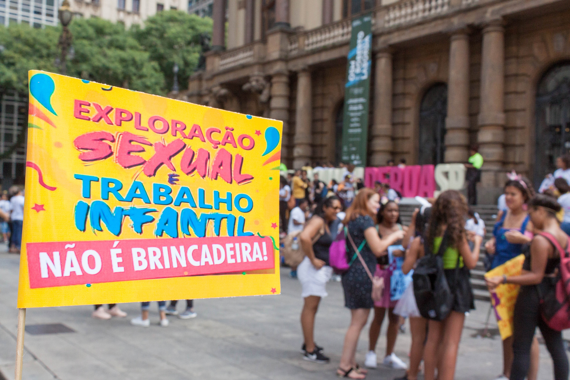 Público concentrado em frente ao Theatro Municipal. Ao lado esquerdo há uma bandeira pequena em fundo amarelo com letras rosa com os dizeres: exploração sexual e trabalho infantil não é brincadeira. 