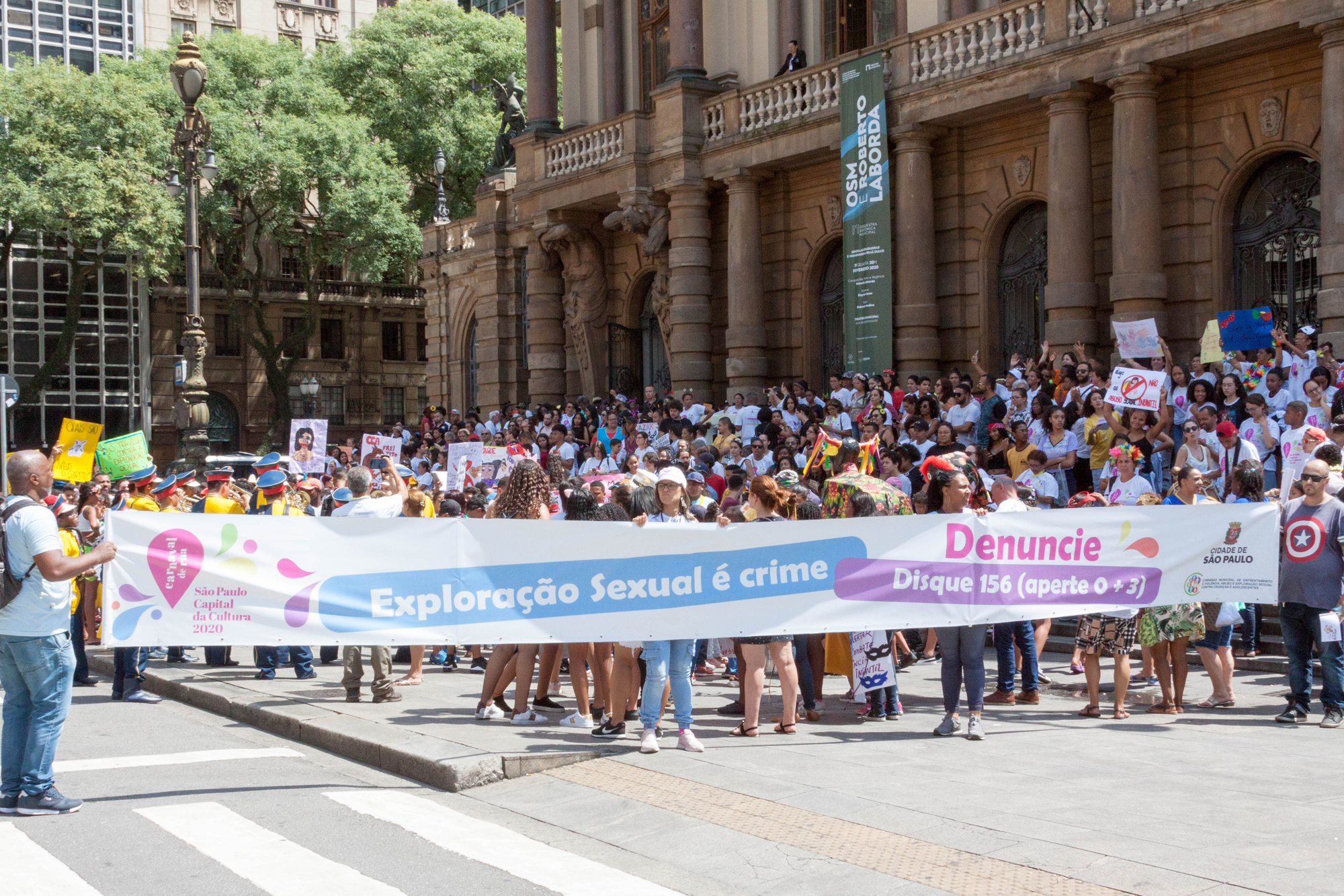 Pessoas seguram a faixa contra a exploração sexual infantil em frente ao Theatro Municipal, ao fundo há várias crianças de serviços conveniados. 