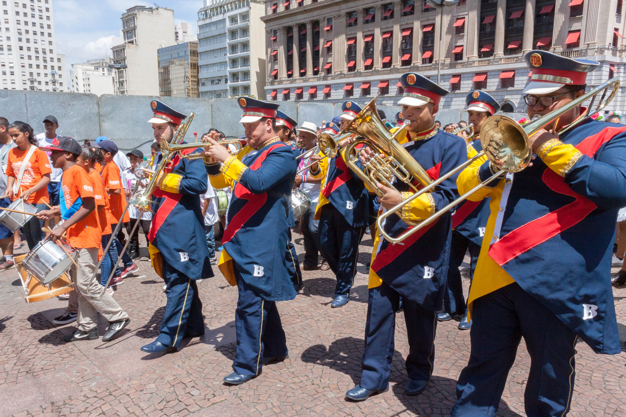 Homens da Banda Marcial Circo Escola Dom Bosco tocam instrumentos, e ao lado esquerdo também há crianças uniformizadas tocando. 