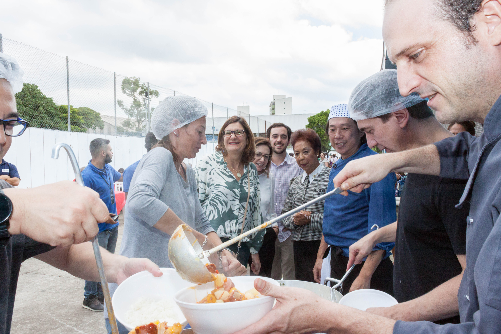 Chefs de cozinha estão servindo alimento em recipiente. Na foto está a secretaria municipal de Assistência e Desenvolvimento Social, Berenice Giannella. 