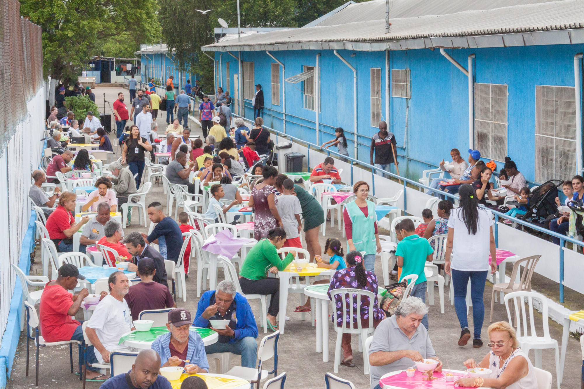 Dezenas de pessoas, entre homens e mulheres, estão sentadas realizando uma refeição ao ar livre.