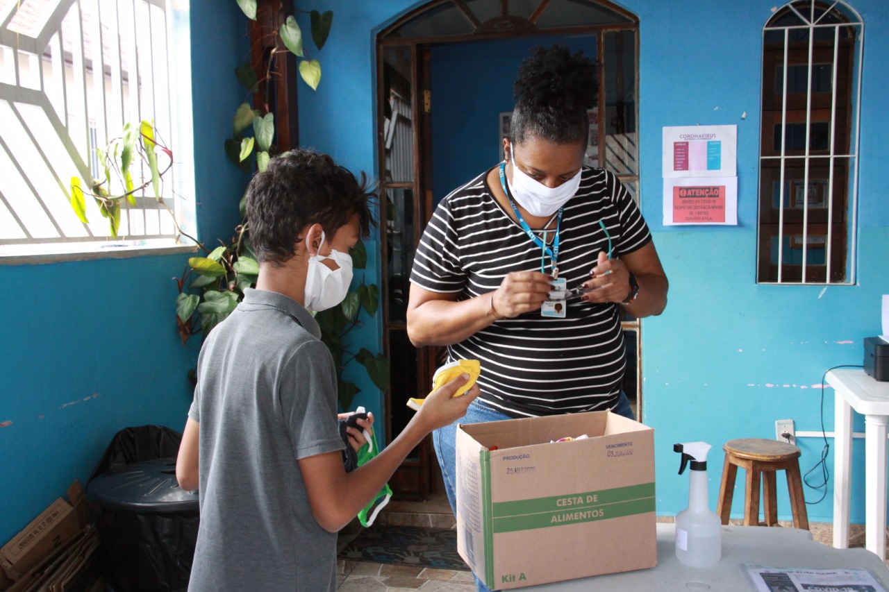Mulher de camisa listrada entrega máscaras de proteção e cesta básica para integrante de família cadastrada.
