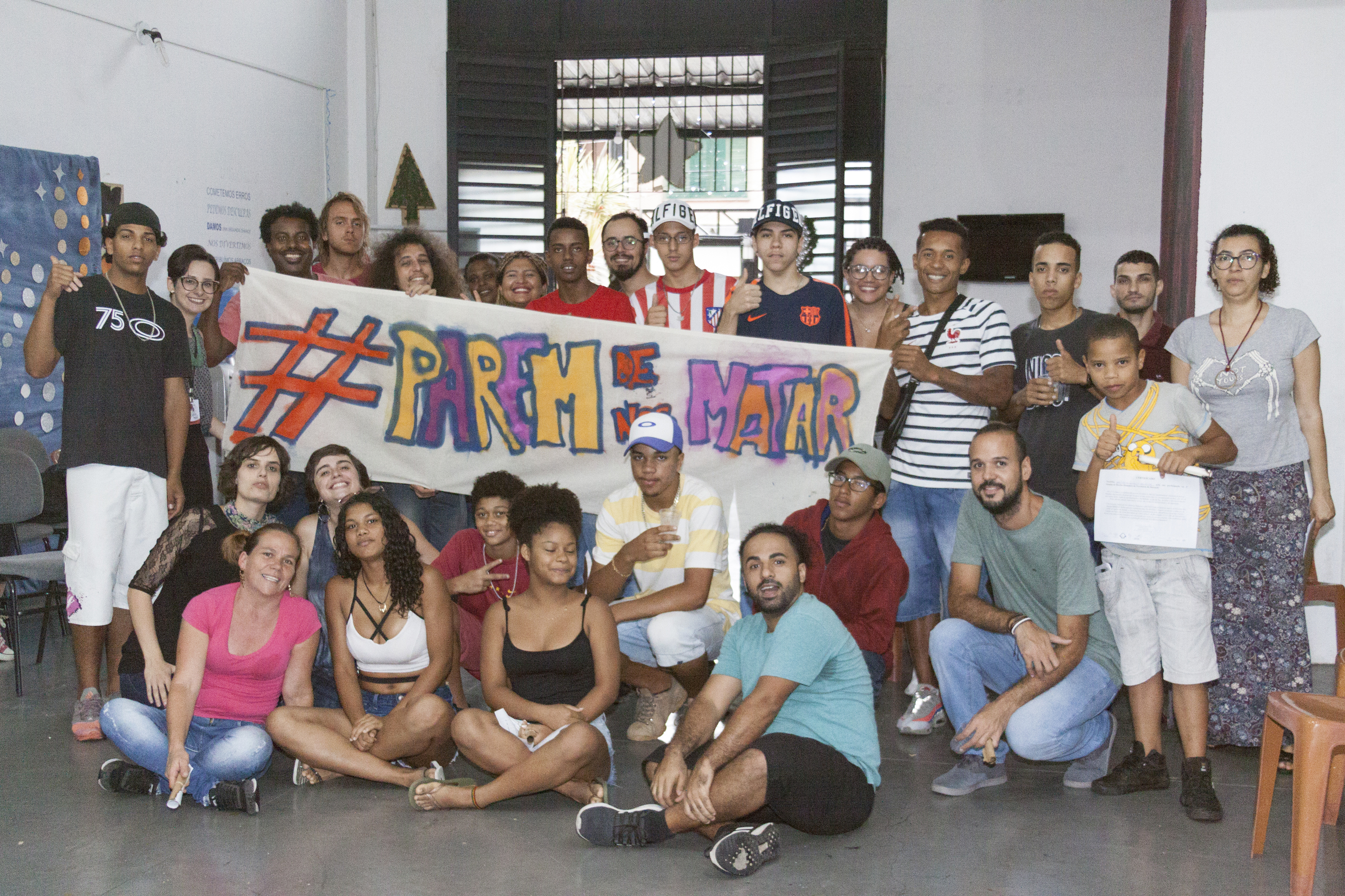 Cerca de 30 pessoas posam para foto com cartaz branco escrito “Parem de nos matar” em letras coloridas. 10 pessoas estão sentadas e as demais estão em pé