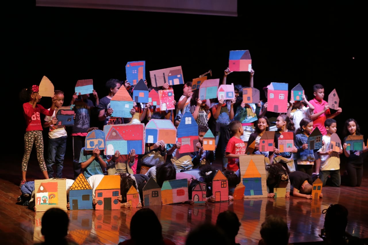 Várias crianças em cima de um palco segurando ilustrações de casas.