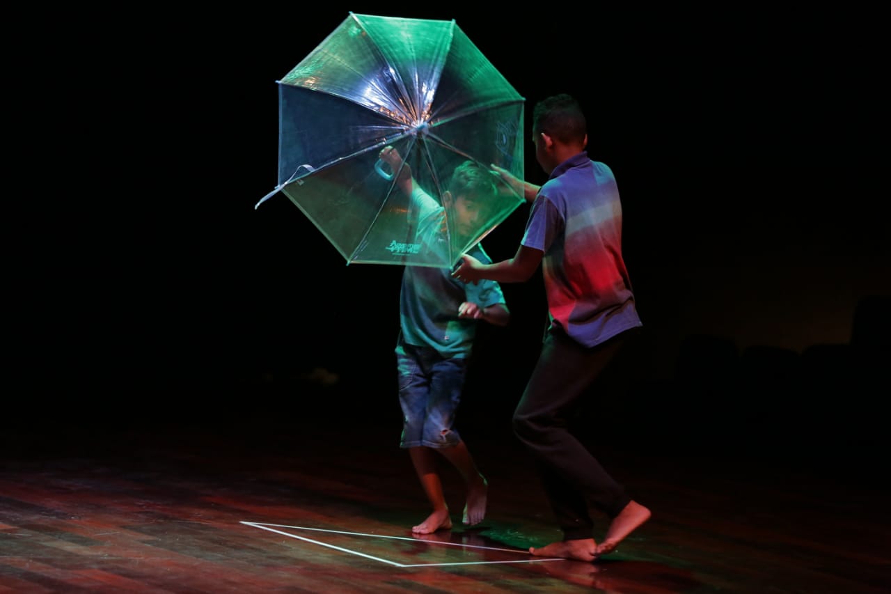 Dois meninos em cima do palco, um deles segura um guarda chuva e eles parecem estar dançando.