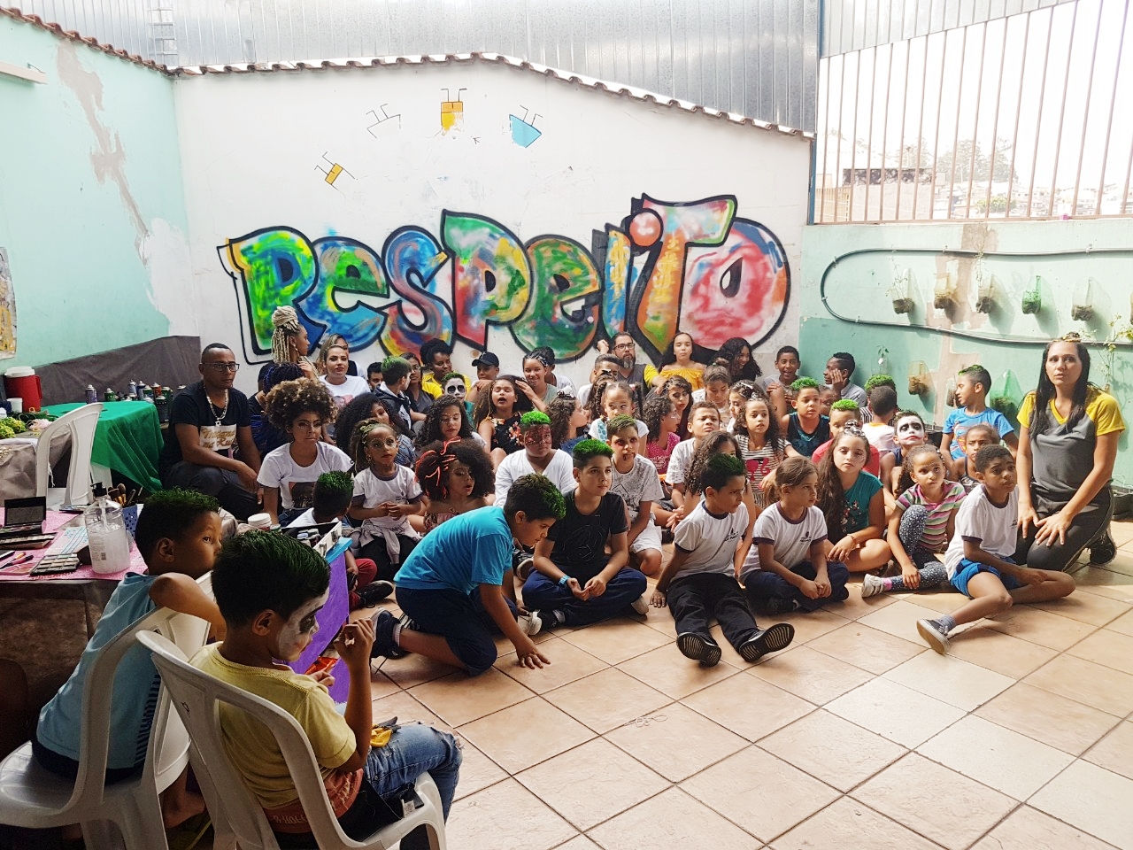 várias crianças posam para a foto sentadas no chão, atrás há uma parede branca em que pintaram a palavra “RESPEITO” de maneira colorida e grande, visando destaque.