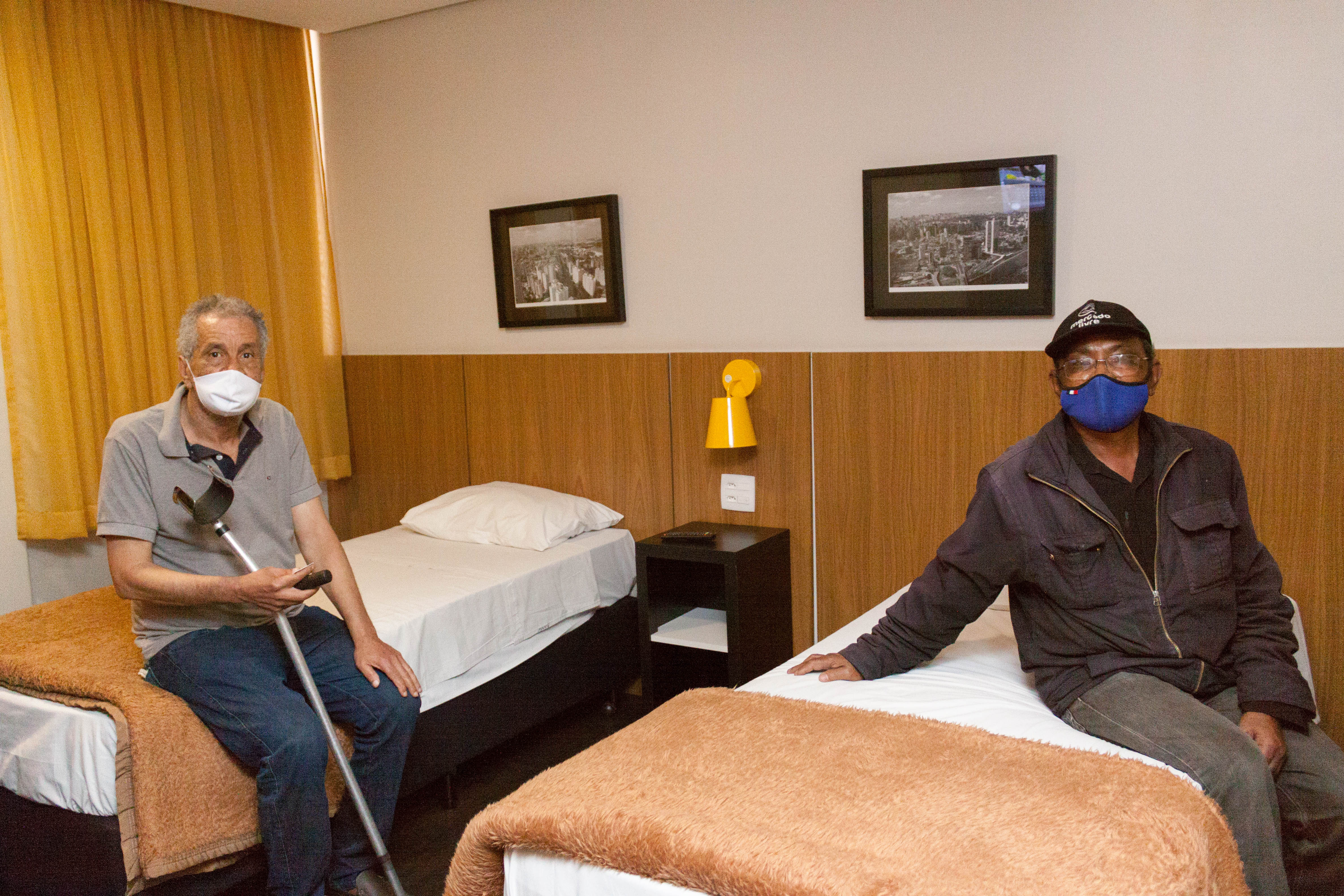 Dois idosos posam para foto. Estão sentados em camas dento do quarto de hotel, que é decorado com quadros da cidade de São Paulo e cores neutras como bege e branco. Ambos usam máscara de proteção.