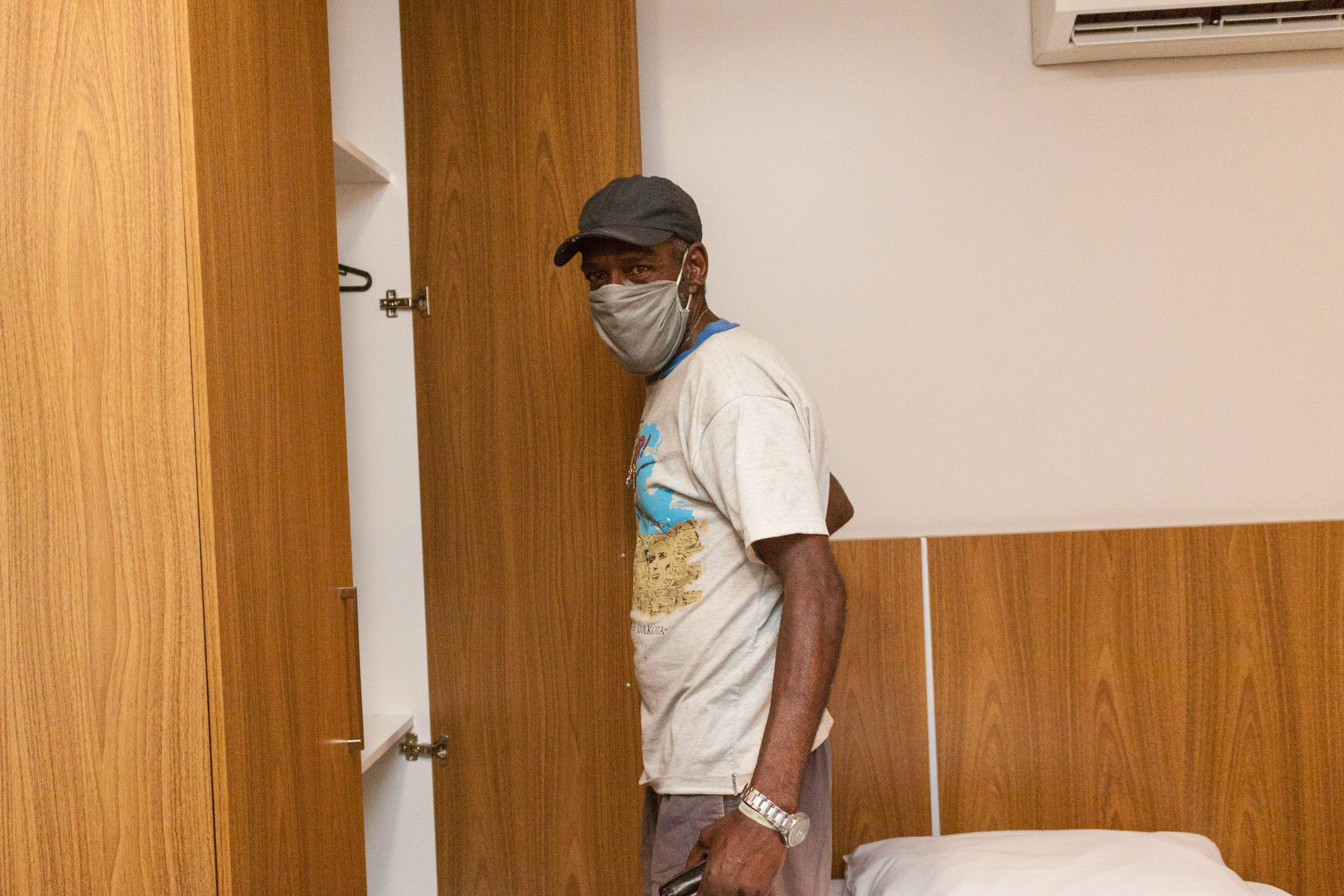 Um idoso de abre a porta do armário para guardar seus pertences. Ele está usando uma camiseta estampada, boné em tom escuro e máscara de proteção.