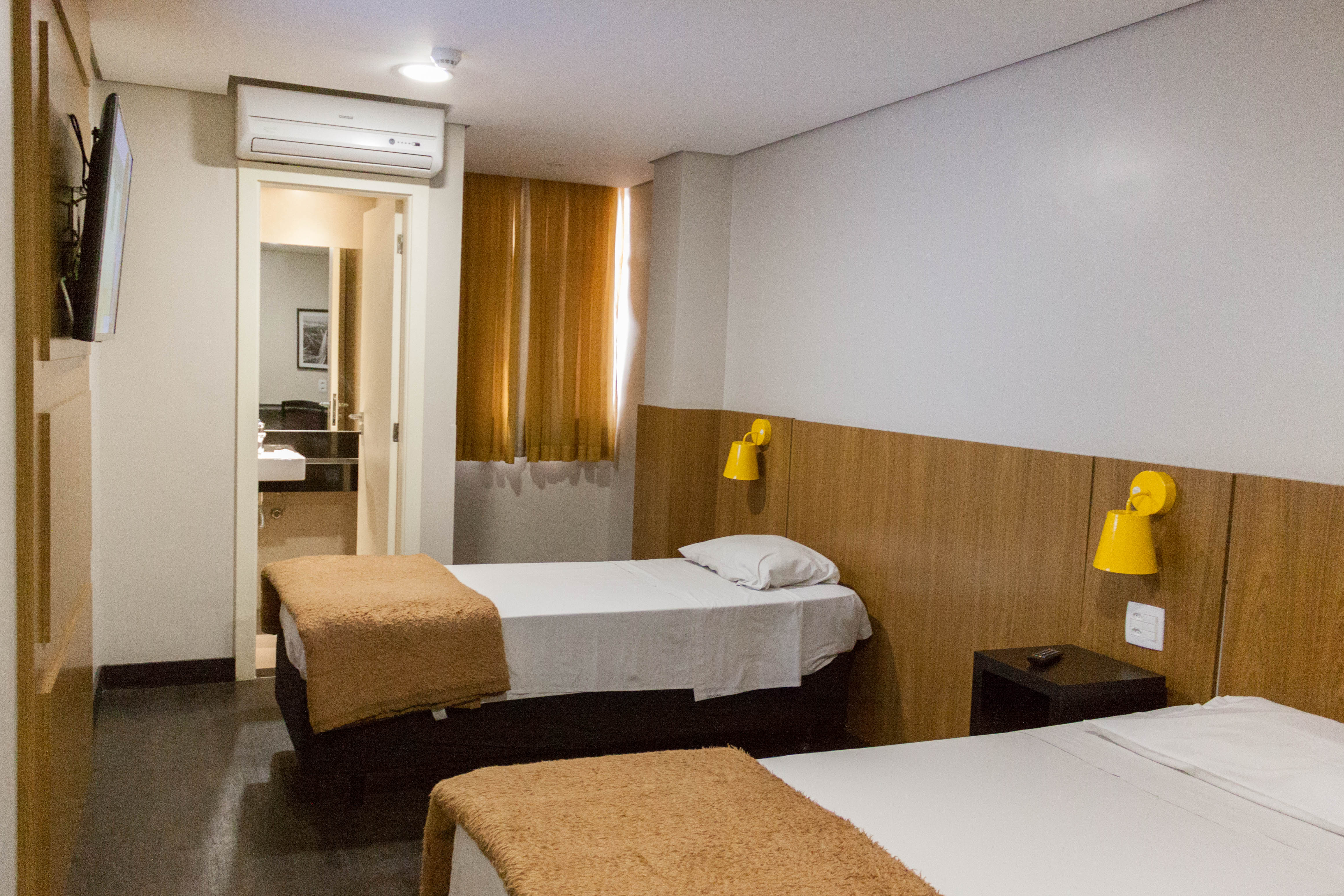 Um quarto com duas camas posicionadas em distância segura. Quadros da cidade de São Paulo e cores neutras compõem o ambiente que possui um banheiro, ar-condicionado e cortinas. 