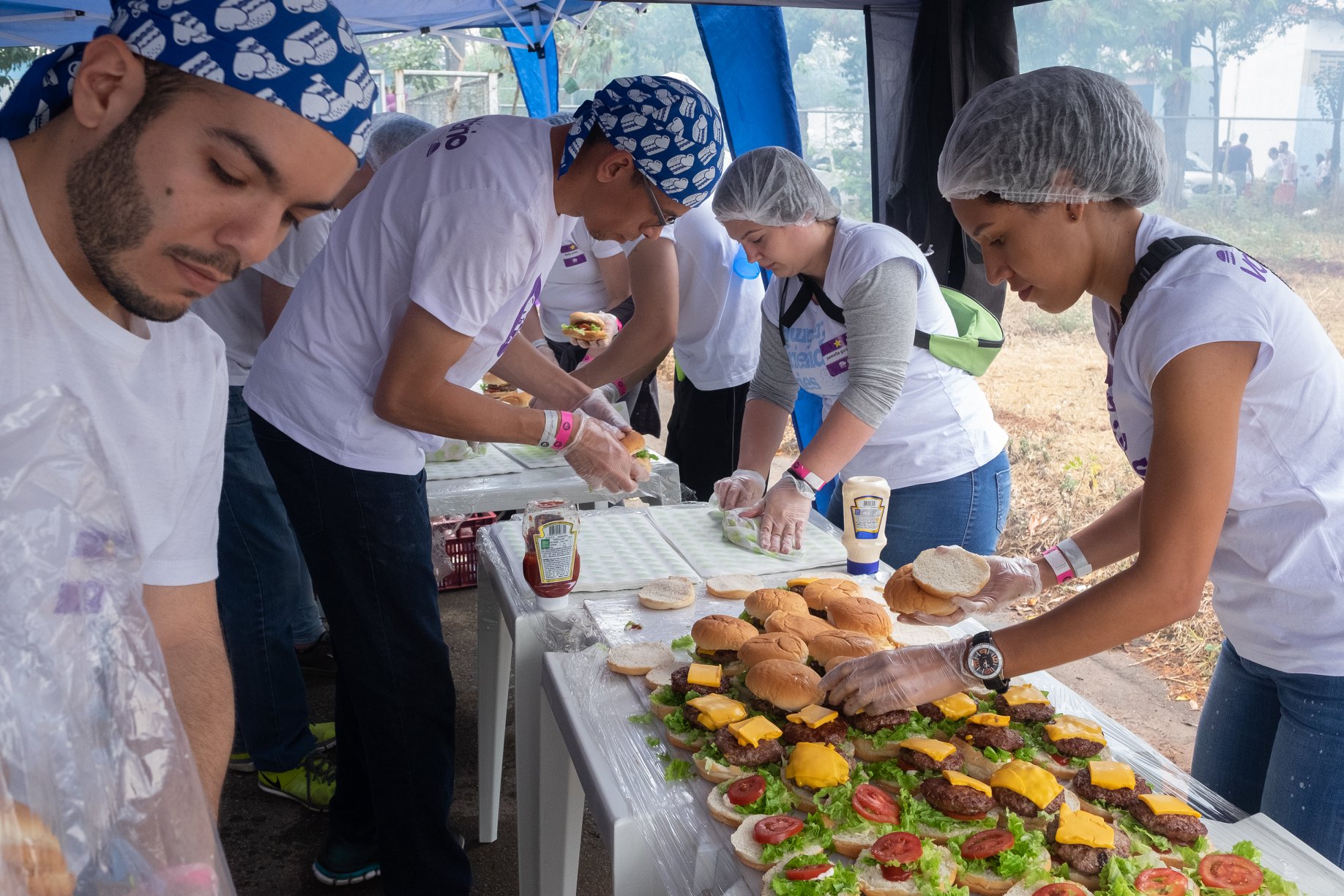 #PraCegoVer1: Quatro voluntários entre homens e mulheres em frente a uma mesa montando hambúrgueres. 