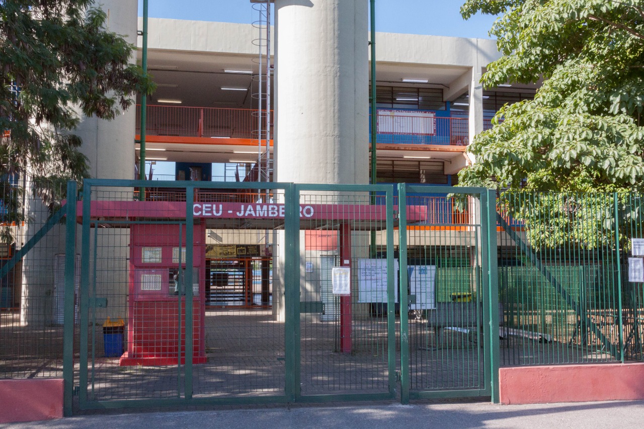 Fachada do CEU Jambeiro com portão verde, estrutura vermelha com letras brancas, duas torres na cor cinza, e de fundo, o prédio com as salas de aulas.
