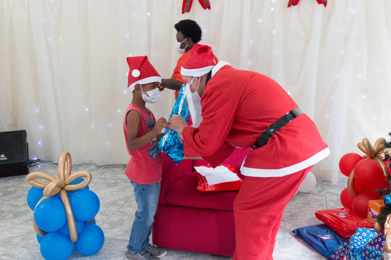 Rubinho Barrichello, vestido com traje de Papai Noel, está à direita da imagem, um pouco encurvado para entregar um presente azul metálico a um menino, um convivente do CCA Marsilac. O menino está à esquerda da foto, vestindo calça jeans, camiseta regata vermelha, chapéu de Papai Noel e máscara branca. A cena se completa com balões a gás azuis, sobrepostos e unidos por laço dourado, fazendo o formato de um presente.