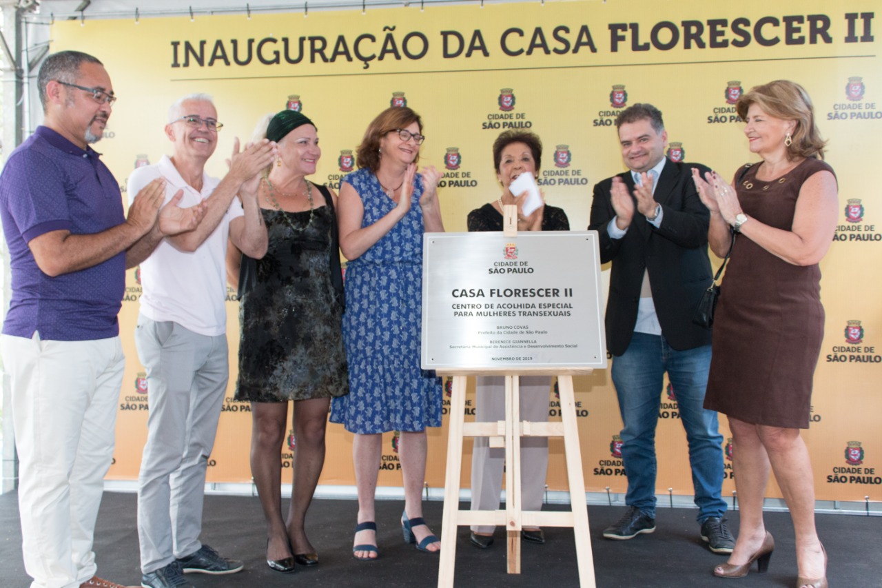 Sete pessoas estão no palco, entre elas a secretária municipal de Assistência e Desenvolvimento Social, Berenice Giannella, ao redor da placa de aço da inauguração da Casa Florescer II.