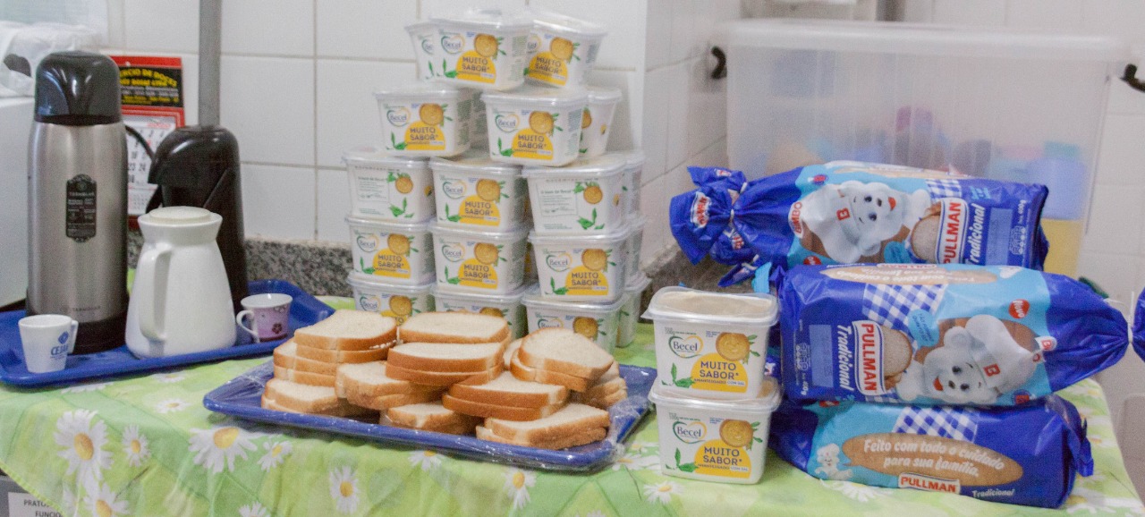 À direita da imagem, três embalagens de pão de forma da Pullman ao lado de 16 embalagens de creme vegetal amanteigado da Becel empilhadas, com lanches dos dois produtos expostos à frente em uma bandeja plástica azul, ao centro da mesa com toalha verde clara. Do lado esquerdo da imagem, uma segunda bandeja de plástico azul com duas garrafas térmicas de diferentes tamanhos e cores, uma cinza e outra preta, uma leiteira de porcelana branca e duas xícaras de porcelana de diferentes tamanhos, uma branca e outra roxa clara, na parte externa. Ao fundo, uma caixa de armazenamento transparente ao lado de uma parede com azulejo branco.